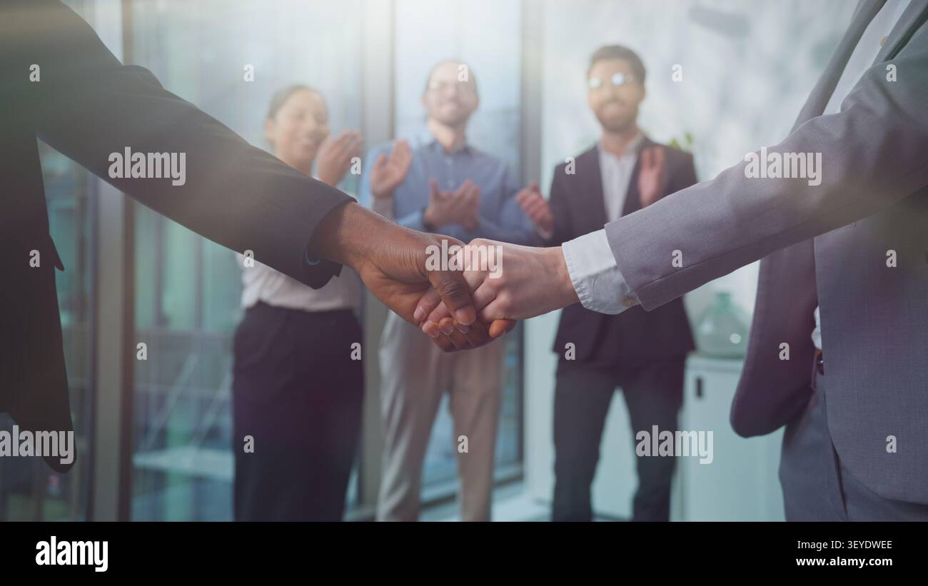 Nahaufnahme kaukasische und afrikanische Geschäftsleute schütteln die Hände in der Nähe von unbeschwerten drei multiethnischen Partnern, die fröhlich im sonnigen Firmenbüro applaudieren Stockfoto