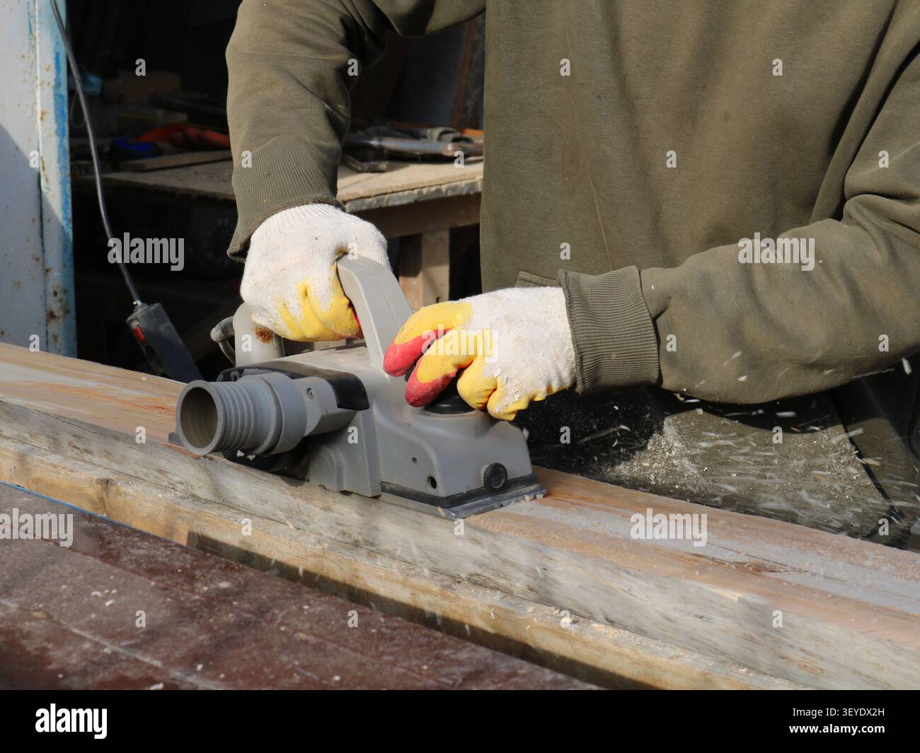 Ein Handwerker mit Handschuhen und khakifarbenem Oberteil hobelt einen Holzbalken mit einem grauen Elektrohobel. Feinspäne fliegen aus dem Werkzeug Stockfoto