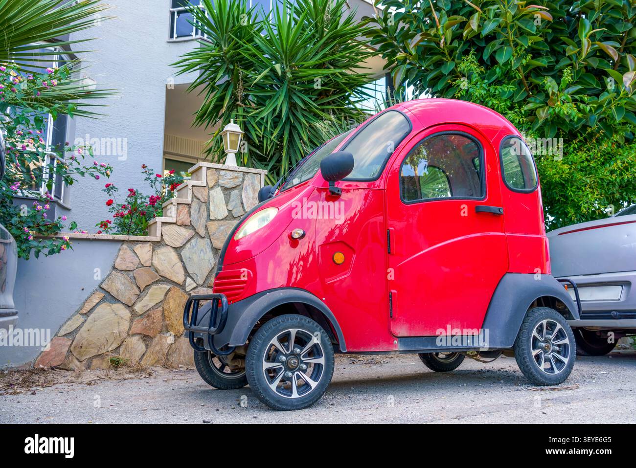 Kompaktes rotes Elektroauto parkt auf einer Schottereinfahrt neben einer Steinmauer mit tropischen Pflanzen und einem Wohngebäude im Hintergrund Stockfoto
