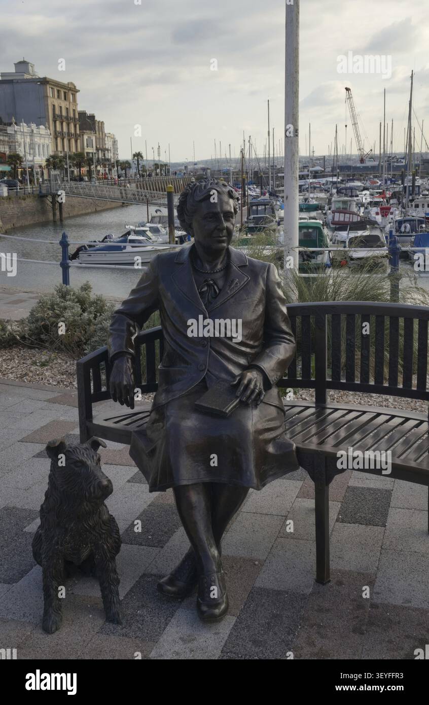 Torquay England - Land: Bronzestatue von Agatha Christie, die auf einer Bank sitzt, mit einem Hund an ihrer Seite an einem Yachthafen mit Booten im Hintergrund Stockfoto