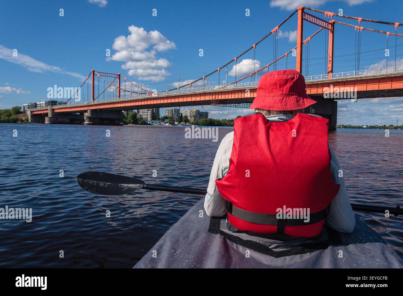 Ein Tourist mit Schwimmweste sitzt in einem Kajak. Er hält ein Paddel und blickt nach vorne. Eine Hängebrücke und mehrstöckige Gebäude sind vor Ihnen zu sehen. Stockfoto