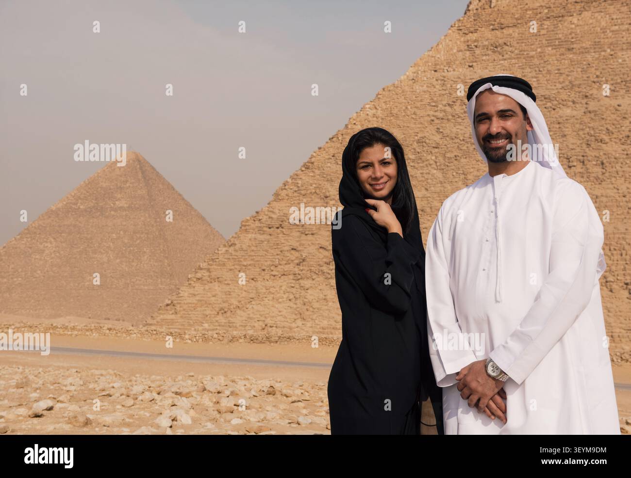 Romantisches Emirati-Paar in traditioneller Abaya und weißem Kandura, das vor den ikonischen Pyramiden von Gizeh warm zusammen lächelt, ideal für MENA-Paare Stockfoto