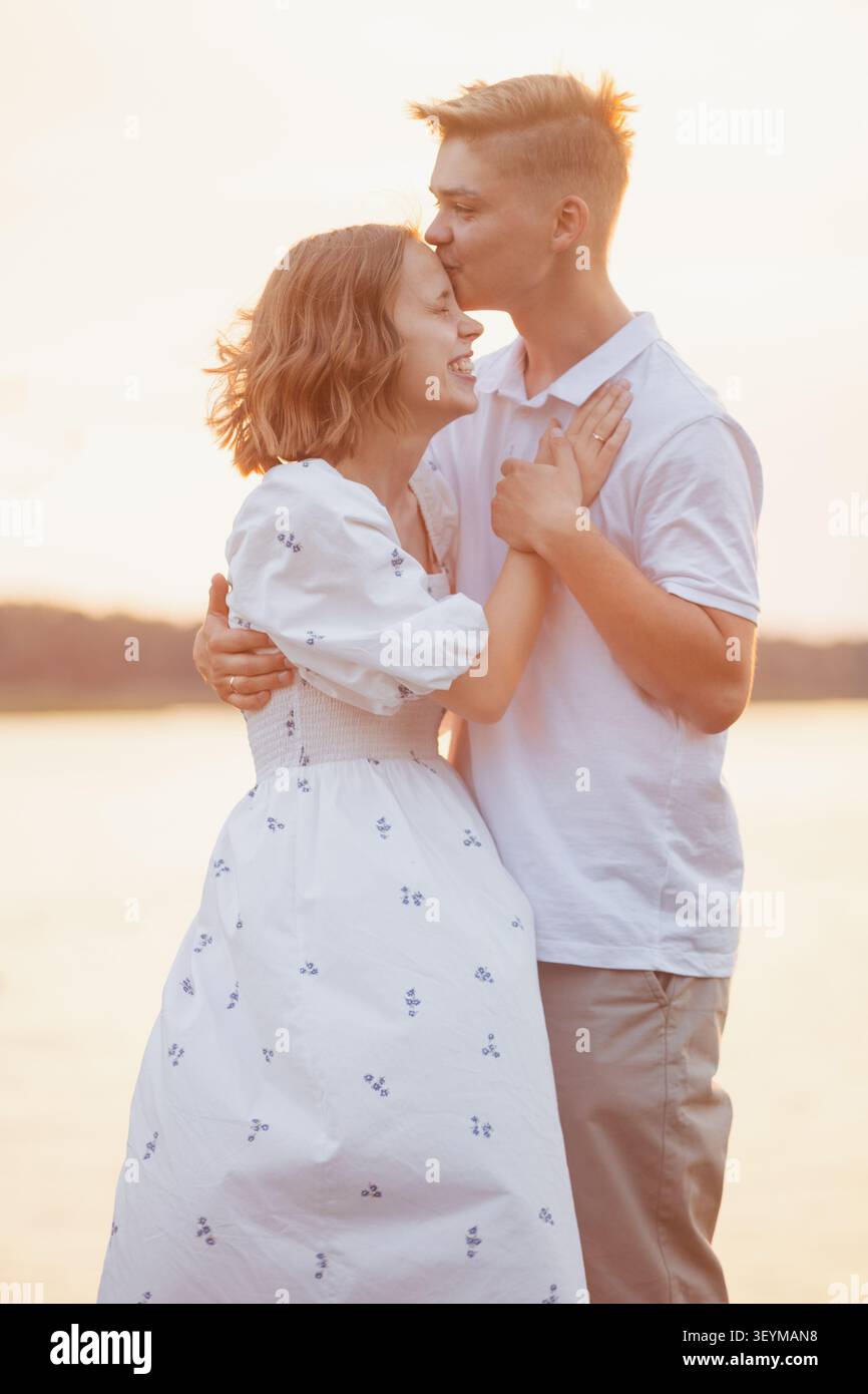 Ein junger Mann küsst seiner lächelnden Freundin auf die Stirn, während sie sich während der goldenen Stunde am Wasser umarmen und eine romantische und freudige Verbindung einfangen Stockfoto