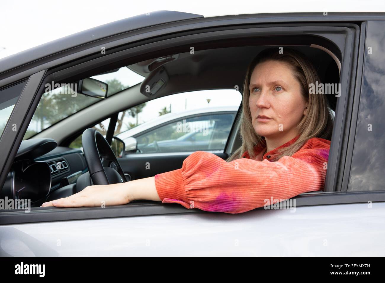 Fahrerin mit blonden Haaren sitzt im Auto auf dem Parkplatz. Sie blickt mit einem fokussierten Ausdruck nach draußen. Horizontal. Nahaufnahme. Stockfoto