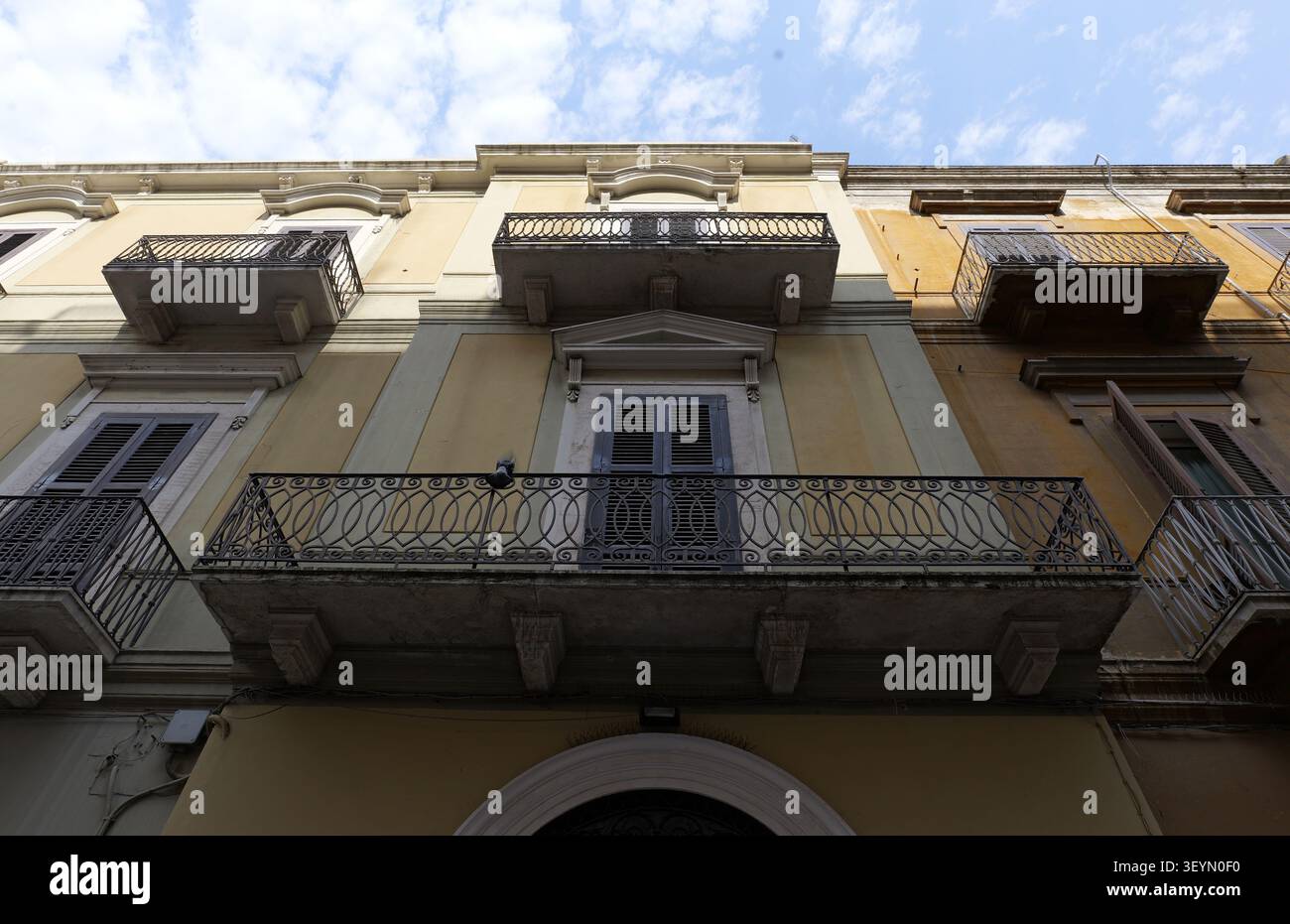 Fassade des Gebäudes mit kunstvoll verzierten schmiedeeisernen Balkonen, geschlossenen Fenstern mit Holzläden und einer gelben, hellen Wand unter blauem, bewölktem Himmel, Showcasi Stockfoto
