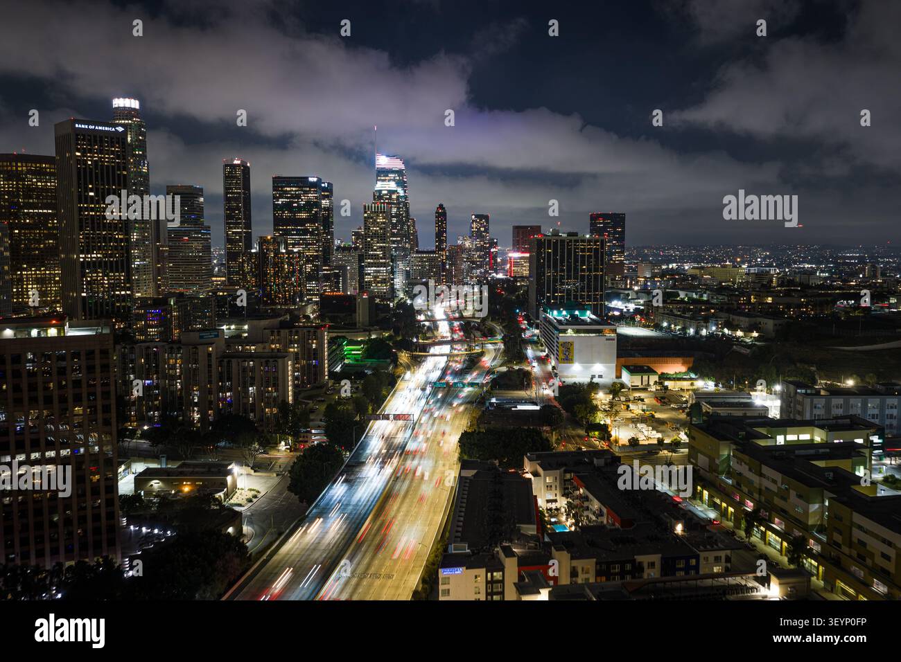 Nächtlicher Blick über die Skyline von Los Angeles mit leuchtenden Hochhäusern, fließenden Lichtpfaden auf der Autobahn, dramatischen Wolken und dichten Stadtgittern Stockfoto