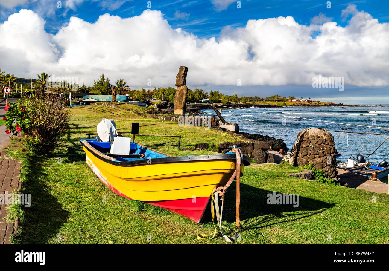 Ein lebhaftes gelbes Boot ruht auf grünem Gras in der Nähe der Küste mit einer historischen Moai-Statue im Hintergrund in Caleta Hanga Roa, Osterinsel, Chile. Stockfoto
