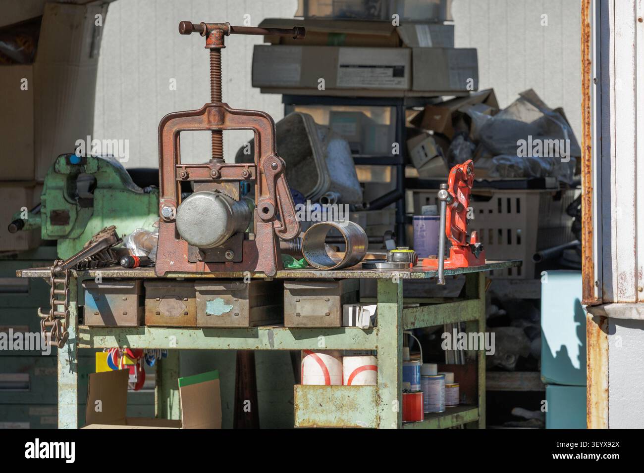 Unordnungige Werkbank mit altem Scharnier-Rohrschraubstock und Schneider, umgeben von Ersatzteilen, Eisenwaren und Werkzeugen für schwere Metallbearbeitung. Stockfoto