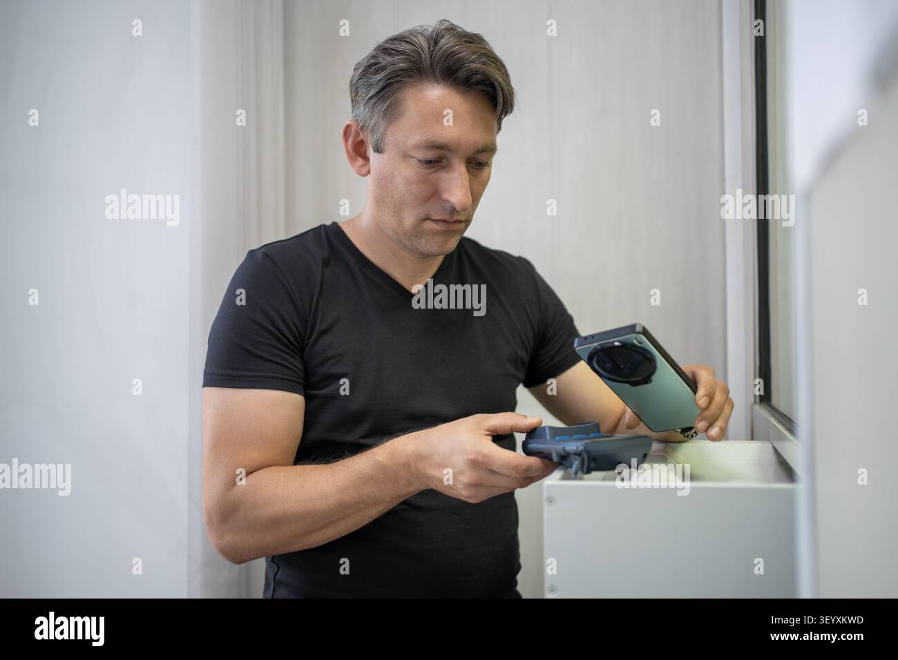 Konzentrierter Mann, der einen Kauf bezahlt, indem er sein Smartphone in die Nähe eines Zahlungsterminals hält. Mobile Wallet, NFC-Transaktion und digitaler Handel. Stockfoto