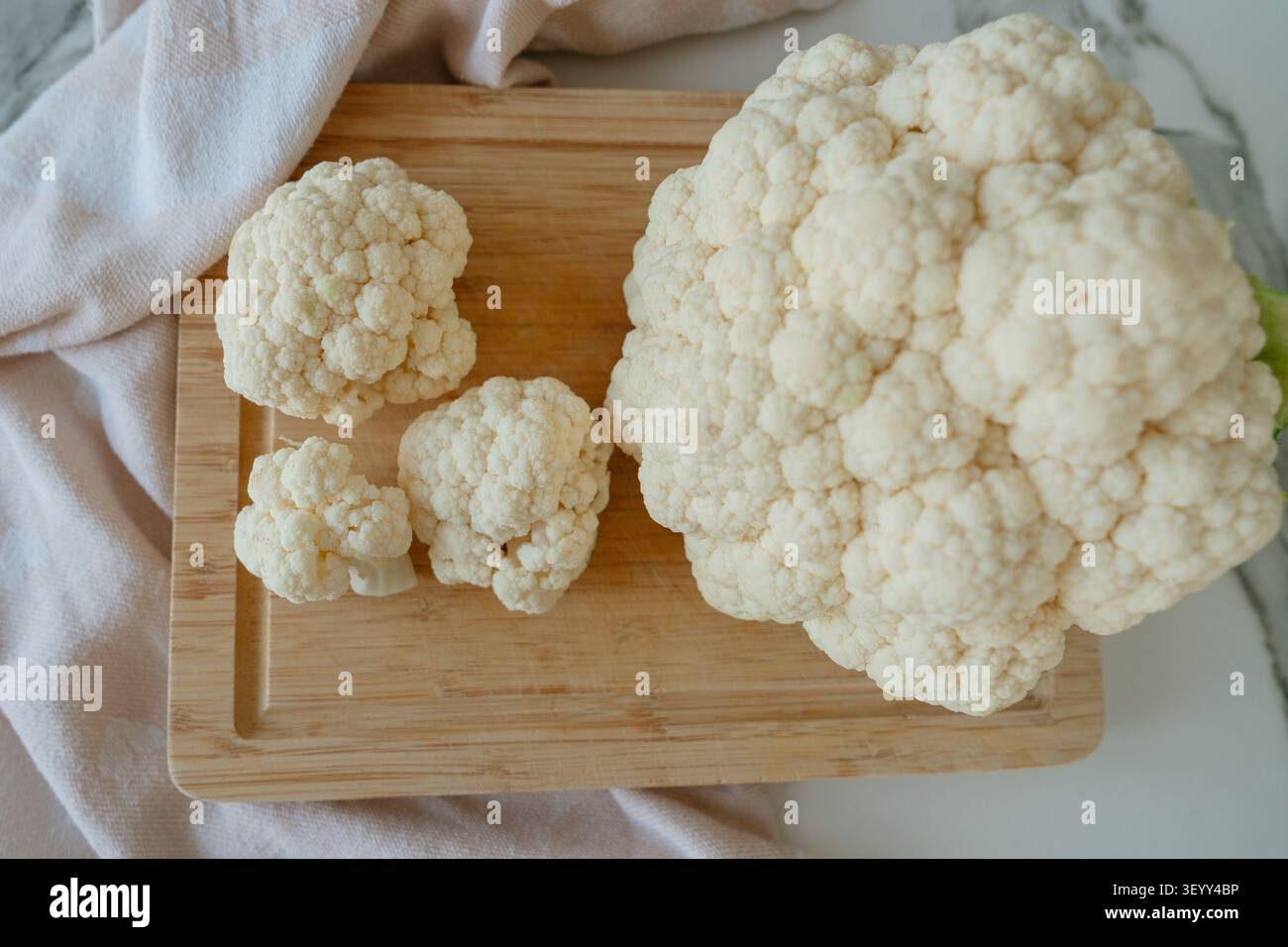 Ein sauberer und frischer Blumenkohl wird in einer Küche auf einem hölzernen Schneidebrett sorgfältig geschnitten, darunter ein Textiltuch. Top-down Stockfoto