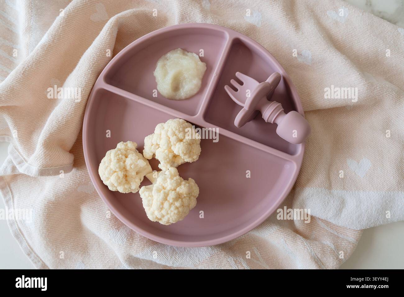 Ein Baby isst Blumenkohlröschen auf einer sauberen Tischdecke mit Herzen, die von oben nach unten aufgenommen werden. Die Einstellung ist rosa und weiß, wobei A hervorgehoben wird Stockfoto