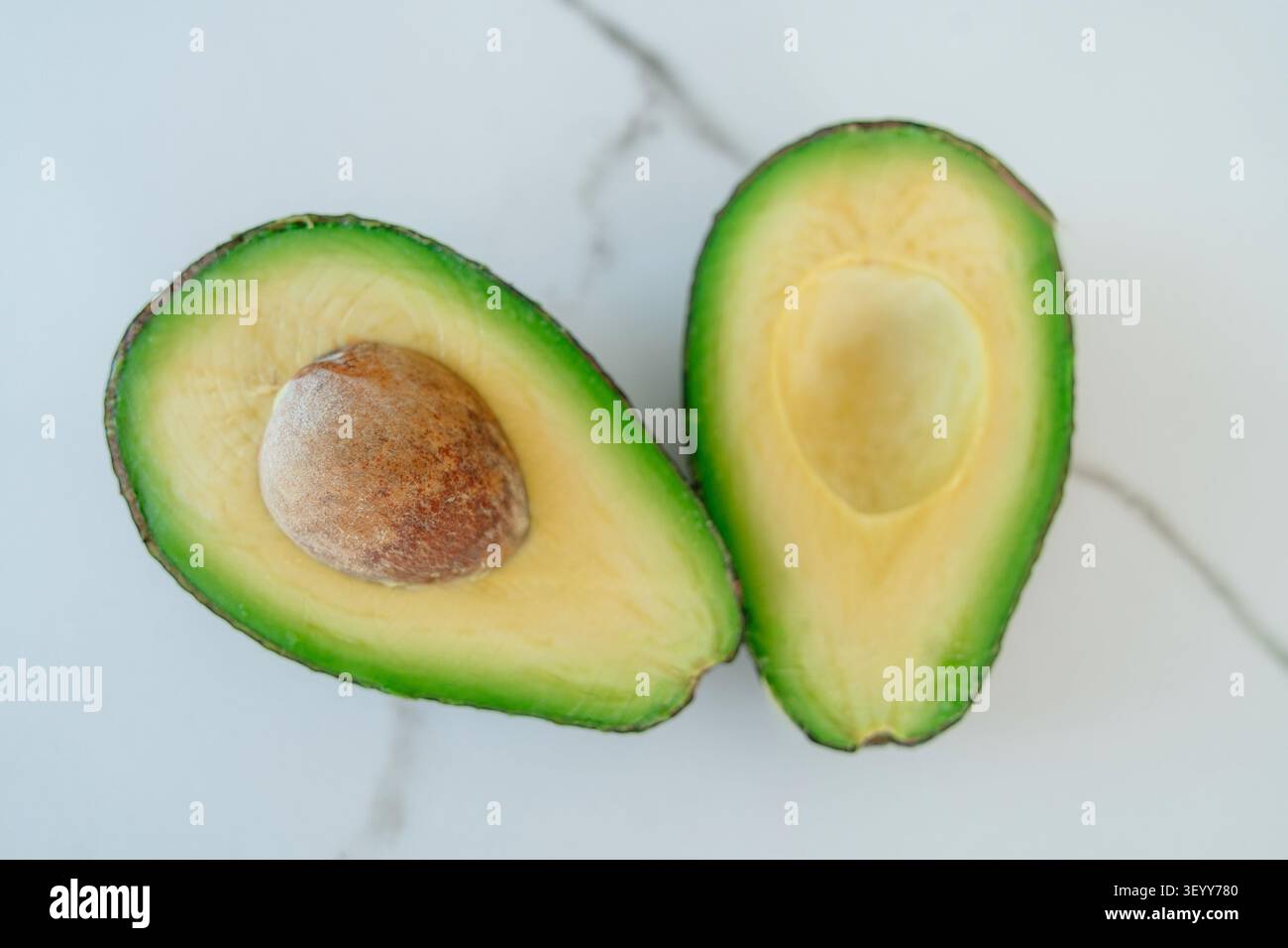 Blick von oben auf eine frische reife Avocado, die in zwei Hälften geschnitten wurde, auf einem sauberen weißen Marmorhintergrund. Eine Hälfte enthält die Grube, die ein gesundes Esskonzept darstellt. Schließen Stockfoto