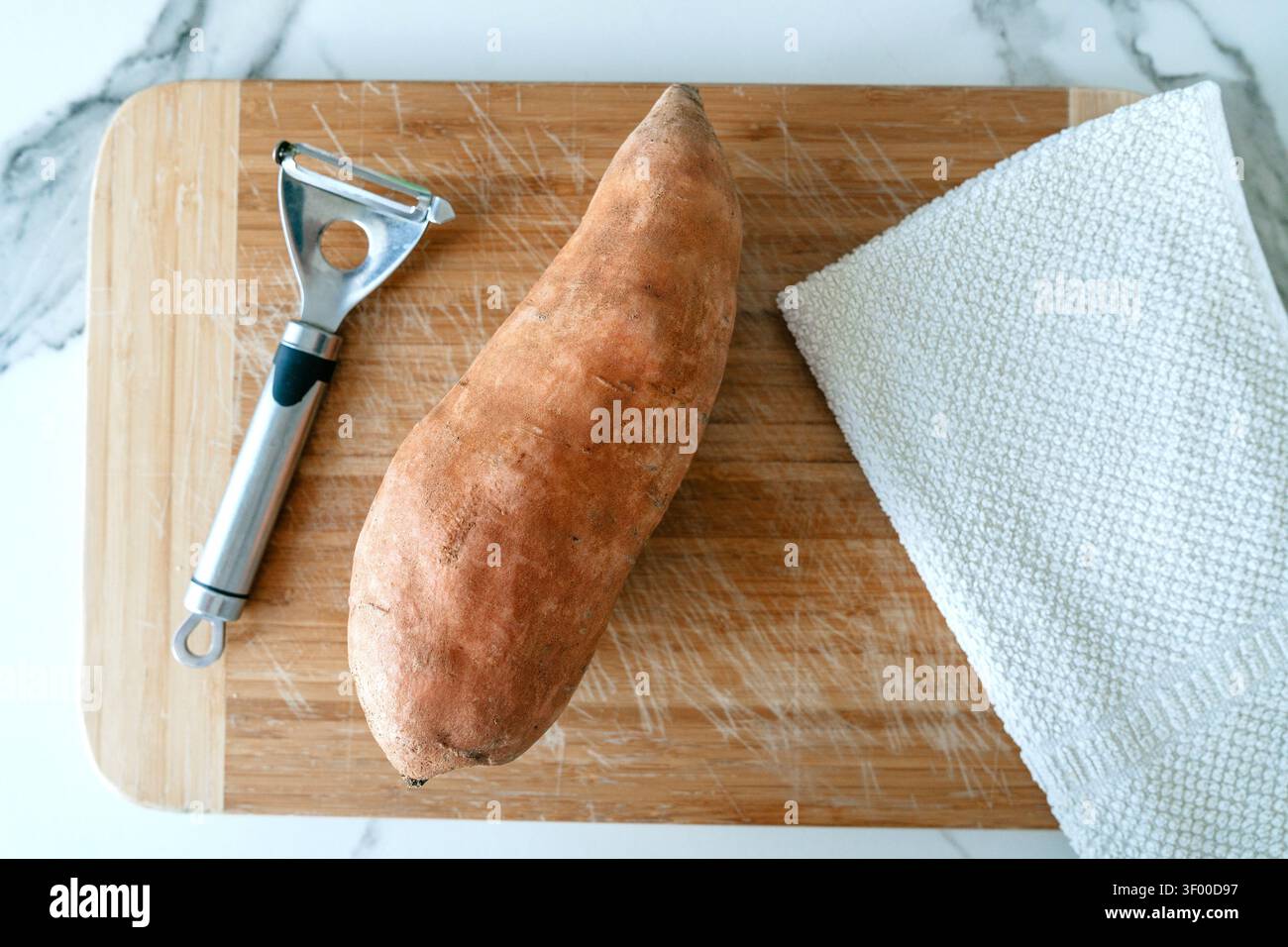 Süßkartoffelschälen, in der Küchenzeile, mit Süßkartoffel, Schäler, Handtuch, mit Holzschneidebrett, Metallschäler, strukturierter Handtuchstruktur, von A Stockfoto