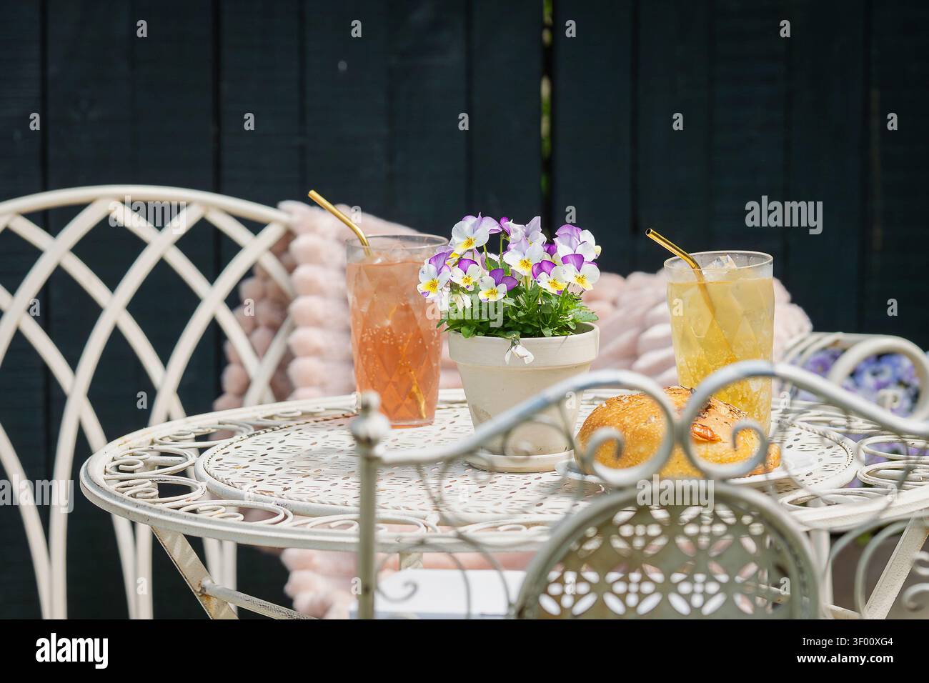 Hausgemachte Rosa- und Zitruslimonade, serviert mit frischem Gebäck auf einem eleganten schmiedeeisernen Gartentisch, dekoriert mit blühenden Stiefmütterchen. Stockfoto