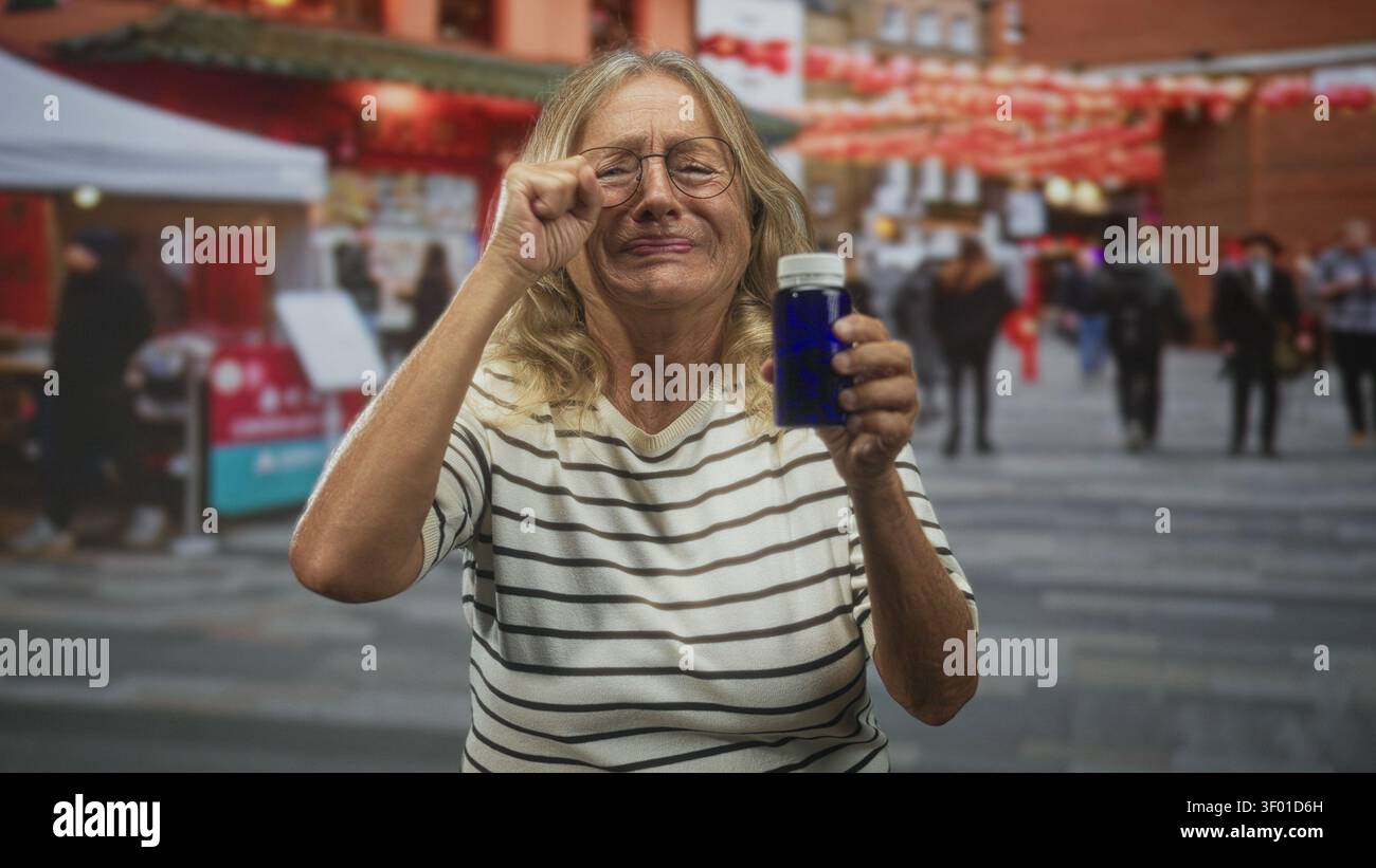 Ältere blonde Frau, die blaue Arzneimittelflasche in der Hand hält, während sie die Augen reibt und auf dem überfüllten Straßenmarkt der Stadt zuschielt; gesundheitliche Bedenken. Stockfoto