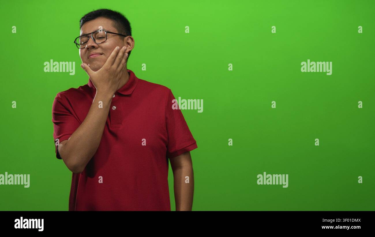 Junger Mann mit Hand auf dem Kinn, der in grünem Hintergrund im Studio posiert, mit rotem Polo und Brille; nachdenkliche Reflexion. Stockfoto