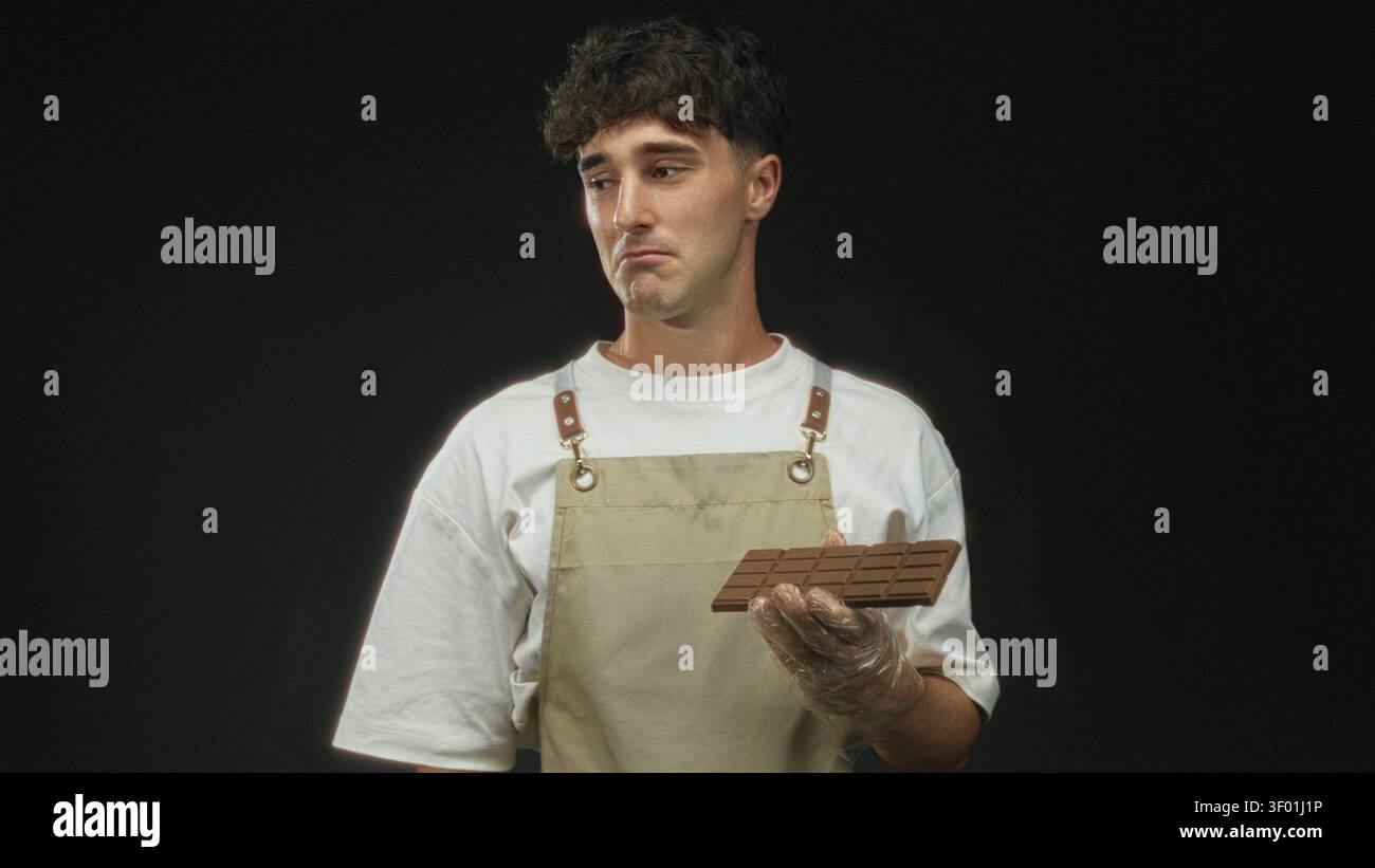 Junger hispanischer Mann, der mit Handschuhen eine Schokoladentafel vor schwarzem Hintergrund hält; ruhige Enttäuschung. Stockfoto