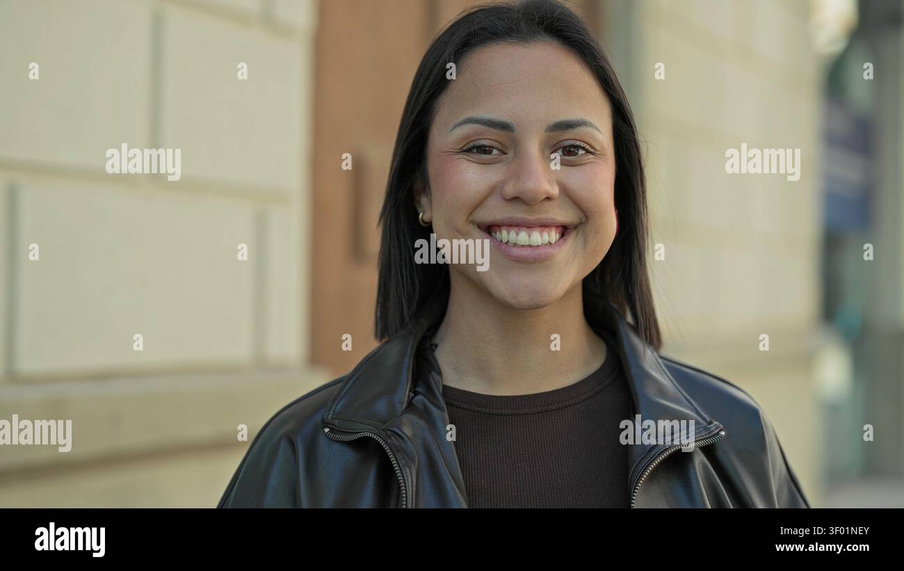 Lächelnde Frau, die Zähne mit einem breiten Lächeln zeigt, Lederjacke und Pullover vor dem Gebäude trägt; Freude am Vertrauen. Stockfoto