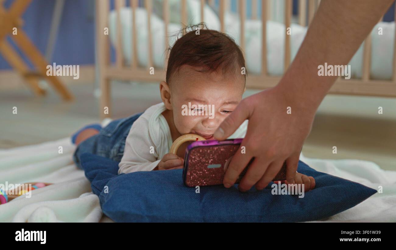 Hispanischer Junge auf dem Bauch, der Holzbeißring hält und im Kinderzimmer mit Krippe und blauem Kissen nach Smartphone greift; elterliche Bindungsneugier. Stockfoto
