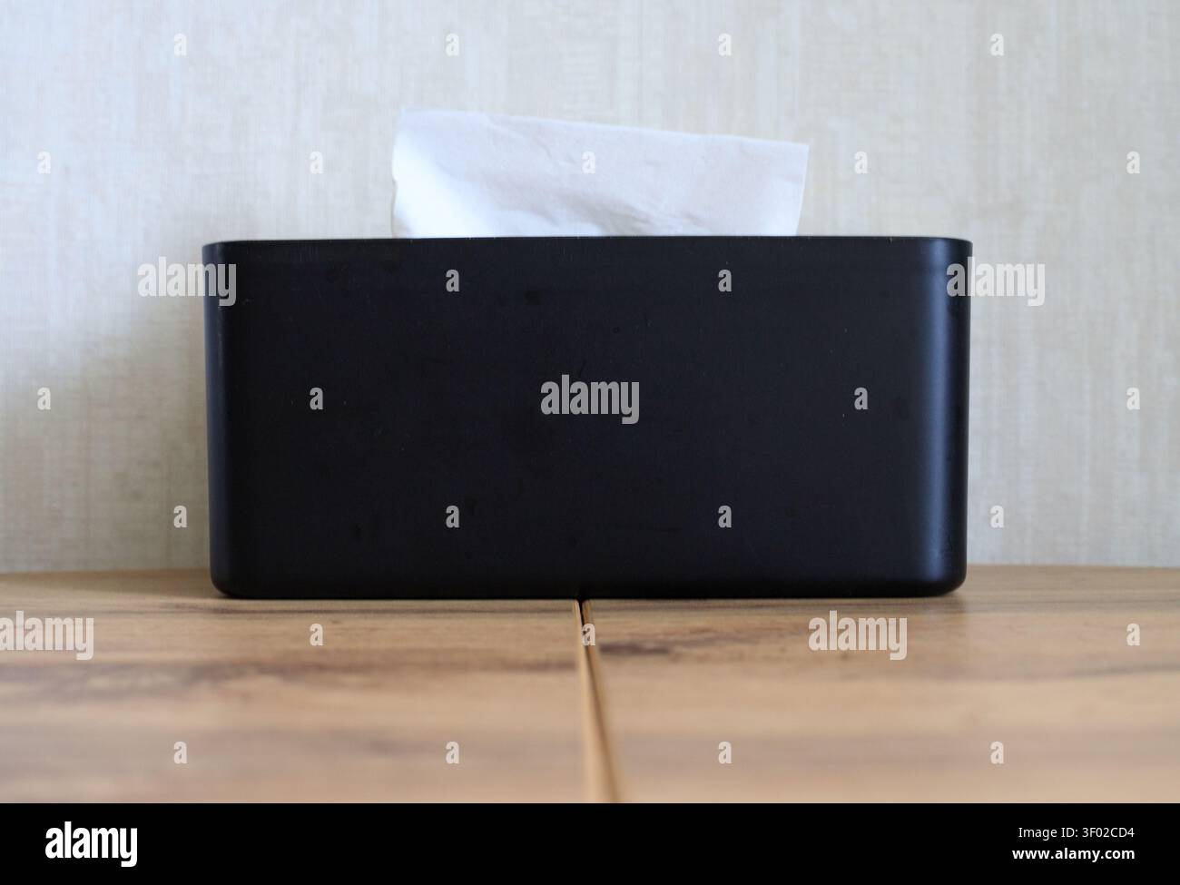 Vorderansicht einer minimalistischen schwarzen rechteckigen Tissue-Box mit einer weißen Papierserviette. Auf einem Holztisch gegen eine helle neutrale Wand gestellt Stockfoto