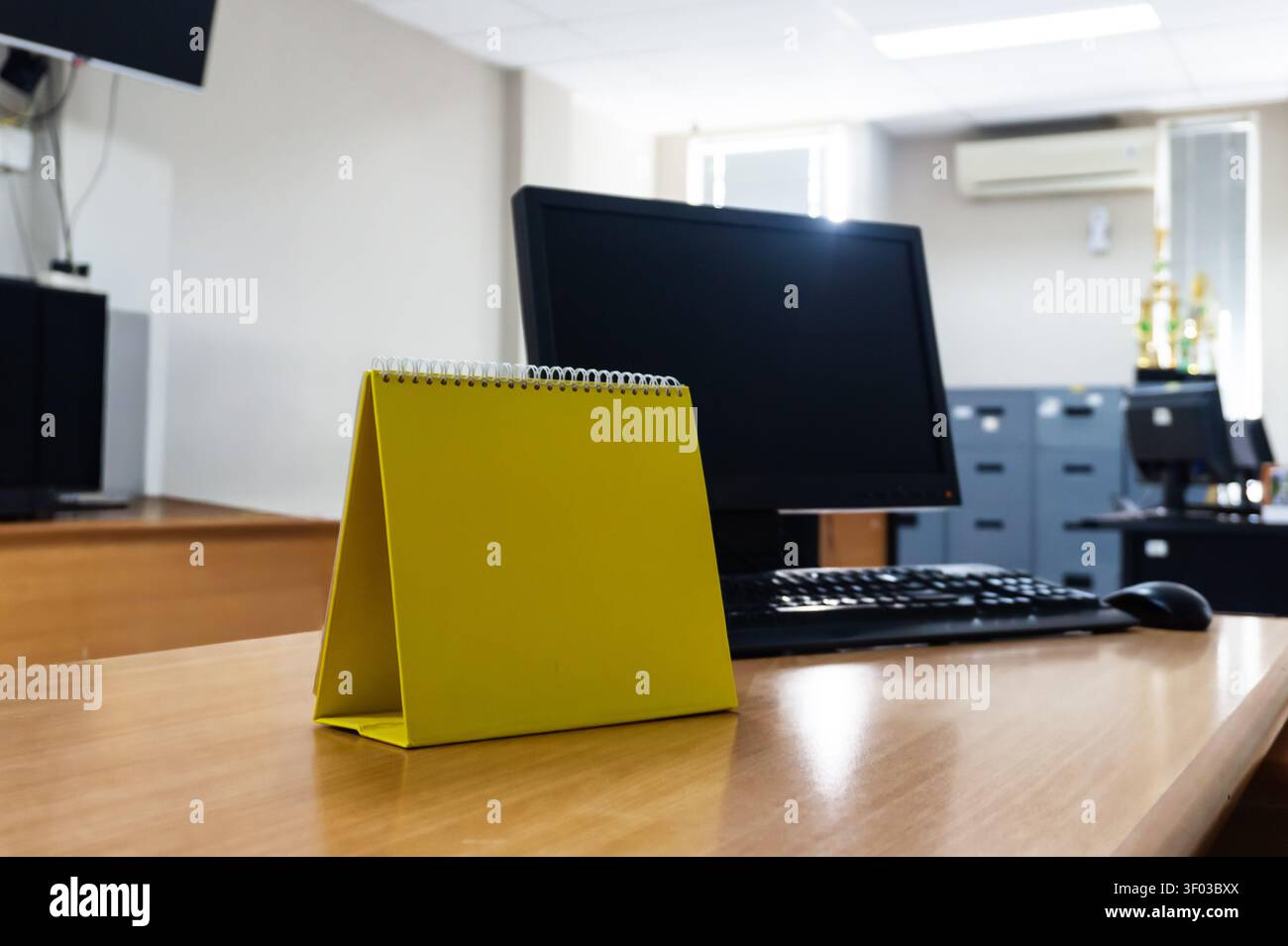 Heller gelber Kalender liegt auf Holzschreibtisch. Schwarzer Monitor und Tastatur in der Nähe, Büroumgebung. Weiche Beleuchtung, ordentliche Einrichtung deutet auf Professionalität hin. Ideal für Geschäft, Planung Stockfoto