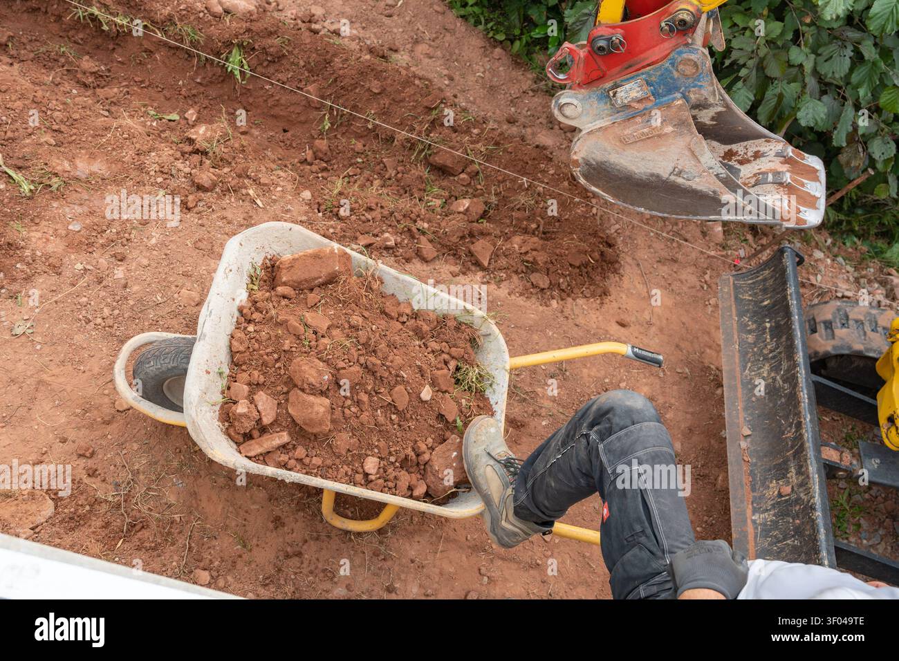 Top-down-Nahaufnahme eines Minibaggerlöffels, der frischen braunen Boden in eine Metallkarre auf einer Baustelle lädt. Hochwertige Bilder für Stockfoto