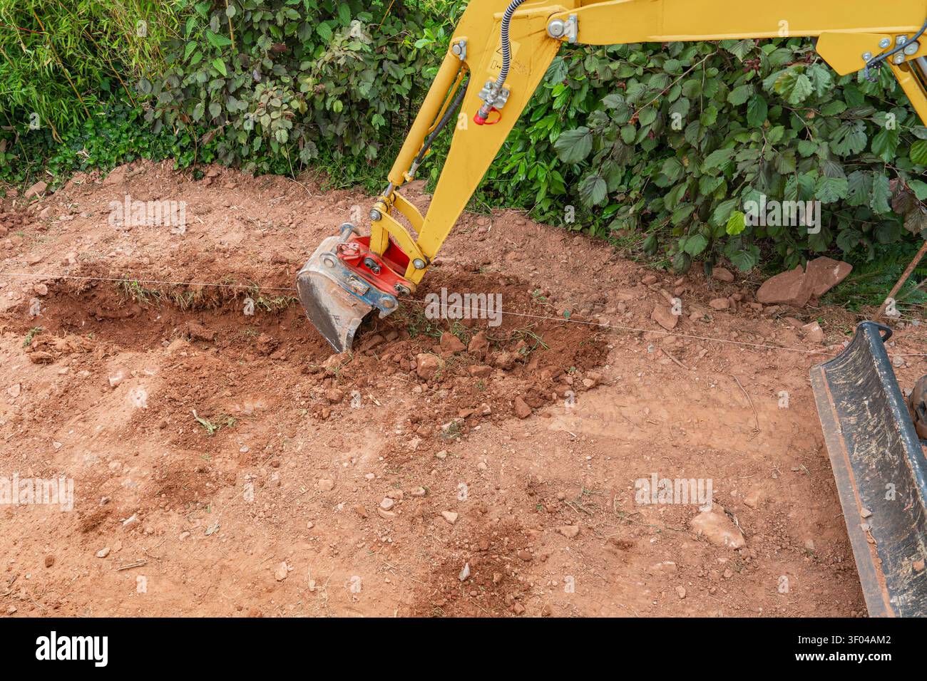 Nahaufnahme eines gelben Minibaggerlöffels, der in trockenen braunen Boden gräbt, um eine Grabenlinie zu bilden. Professionelle Erd- und Landschaftsbauarbeiten an einem Stockfoto