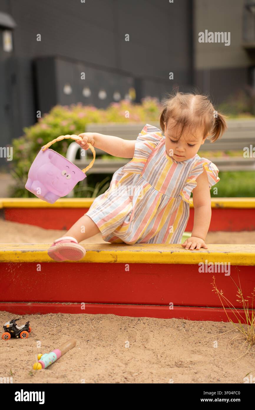 Ein Mädchen in einem pastellfarbenen Sommerkleid, das am Rand einer rot-gelben Sandbox balanciert und einen pinkfarbenen Plastikeimer hält, fokussiert und verspielt i Stockfoto