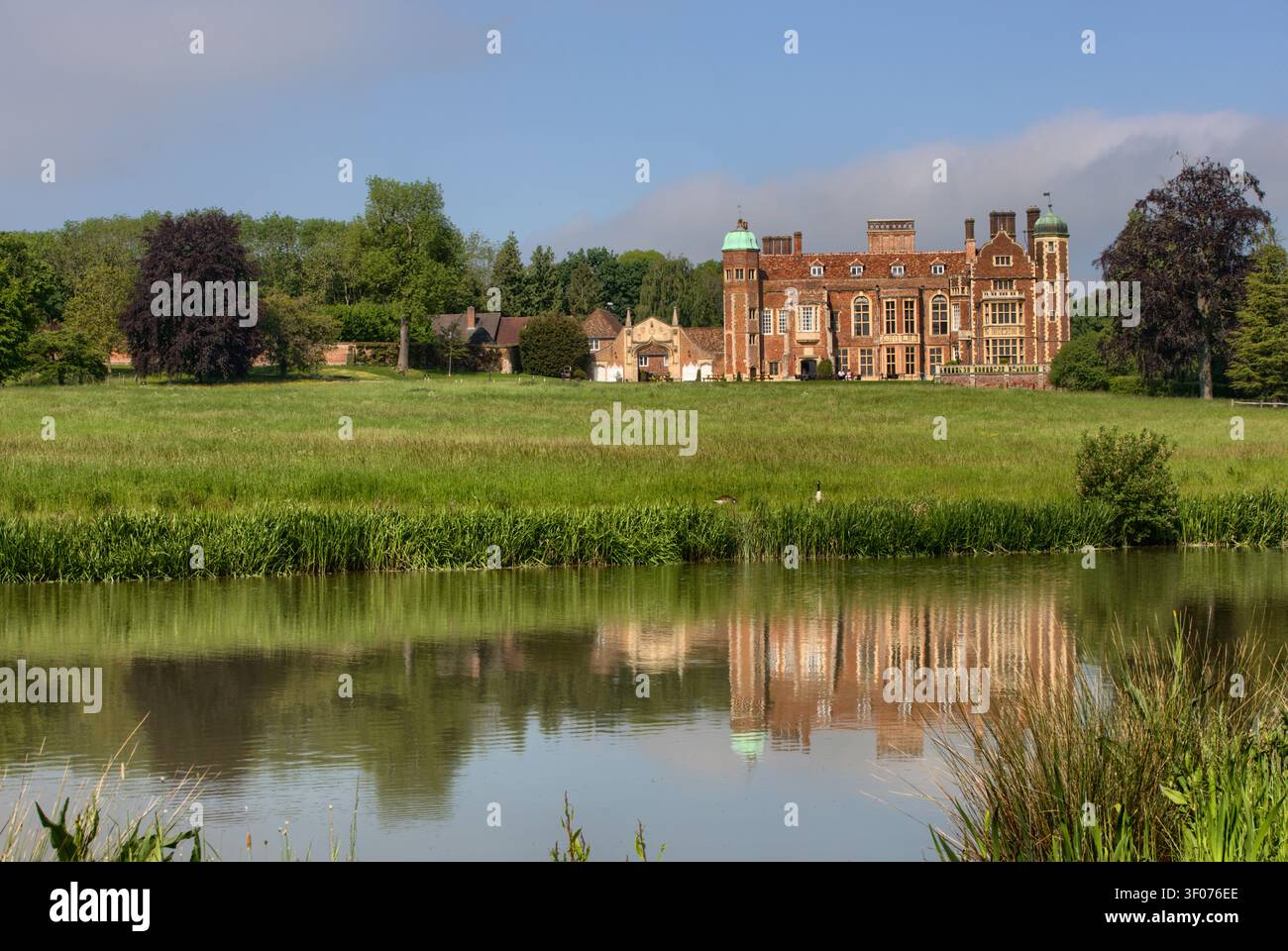Das historische Madingley Hall aus dem 16. Jahrhundert spiegelt sich wunderschön in den ruhigen Gewässern des Sees an einem klaren Sommertag in Cambridge, England, wider. Stockfoto