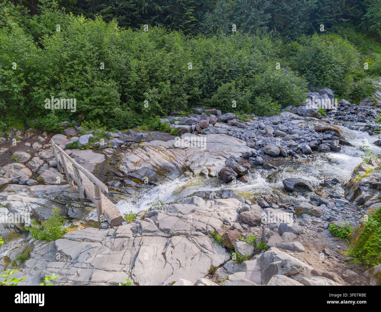 Ein schnell fließender Gebirgsbach kaskadiert über graue Granitfelsen und eine kleine hölzerne Fußgängerbrücke in einem üppig grünen Wald im Bundesstaat Washington. Stockfoto