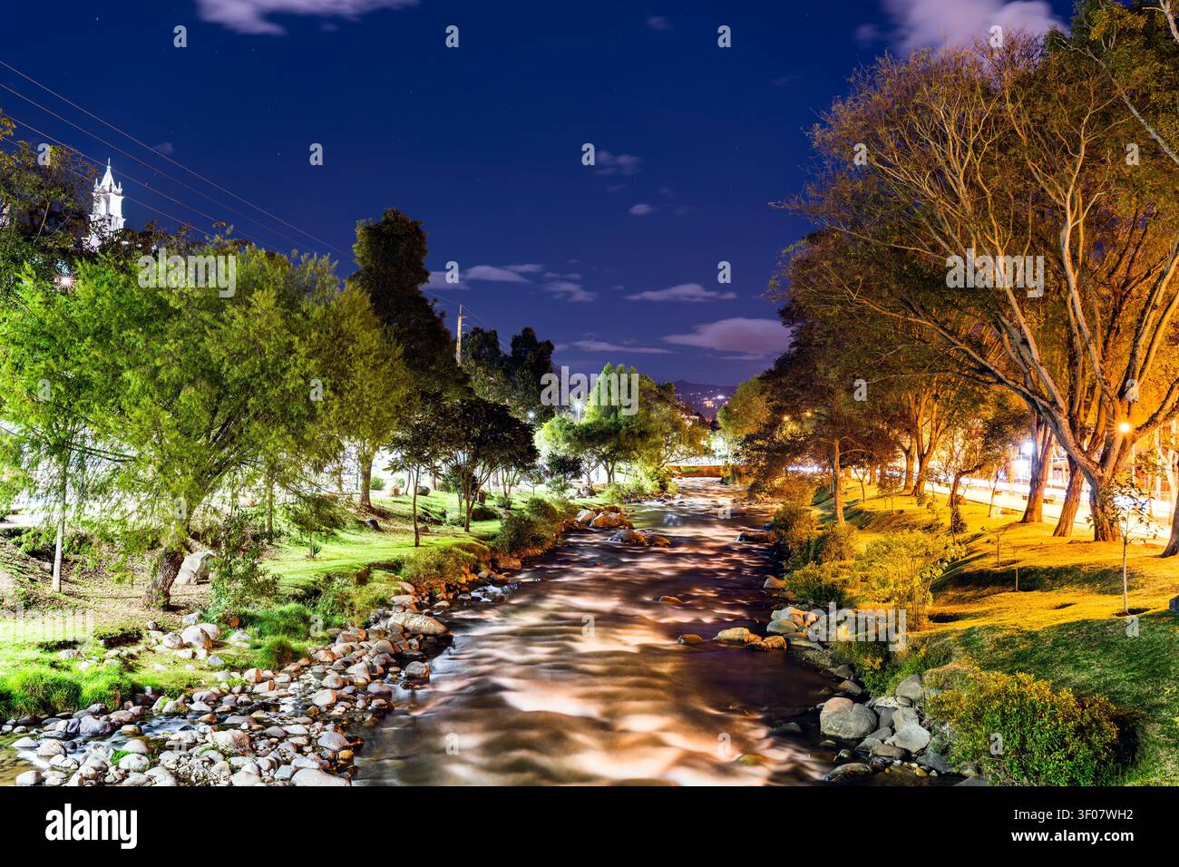Wunderschöne Langzeitausstrahlung bei Nacht auf den Tomebamba Fluss, der durch Cuenca, Ecuador fließt, umgeben von üppigen Bäumen und Lichtern der Stadt. Stockfoto