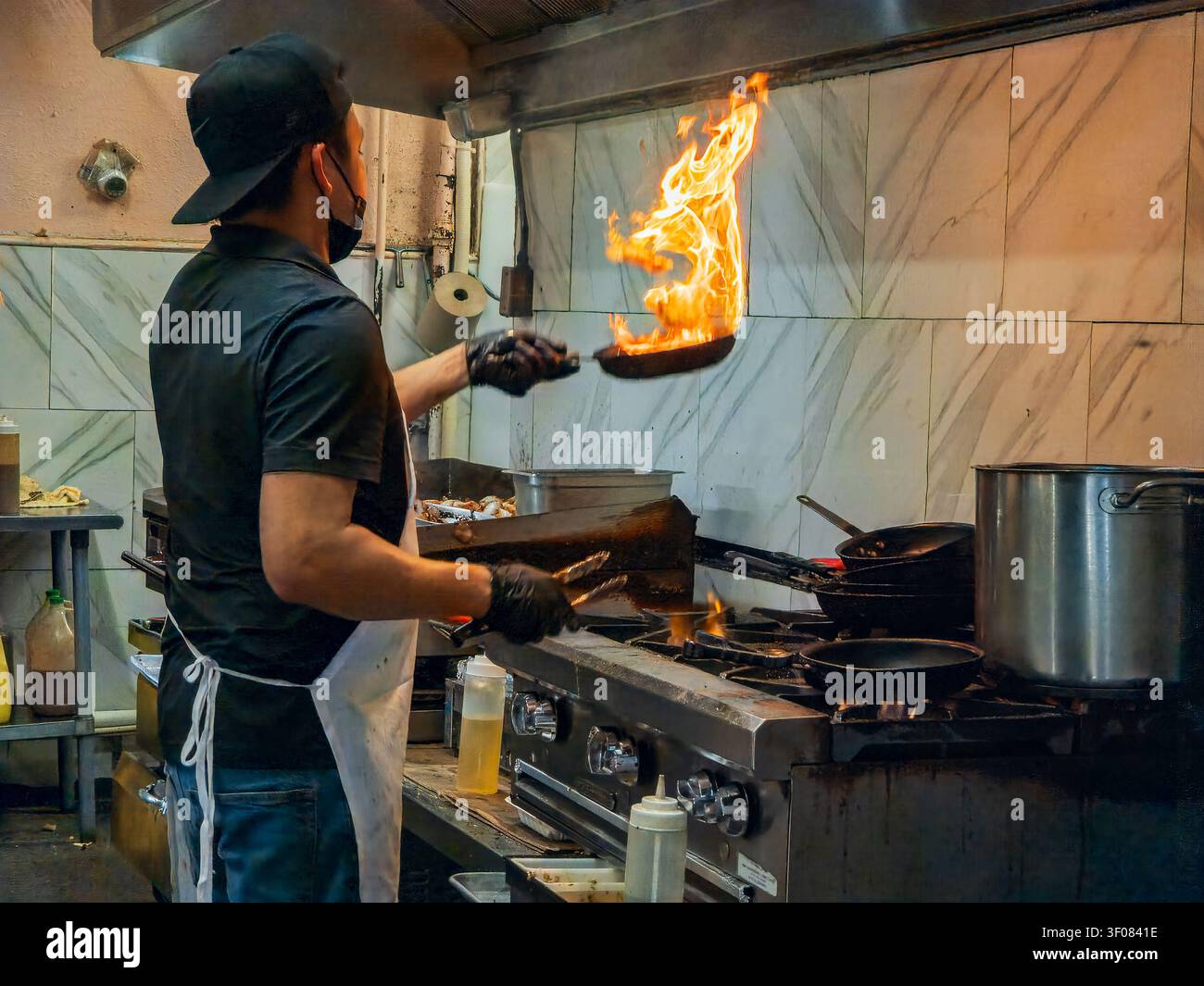 Washington Staat, Vereinigte Staaten, JUN 21, 2026 - Ein Koch mit schwarzer Mütze und Handschuhen wirft eine Pfanne mit hohen Flammen in einer gewerblichen Küche mit Stockfoto