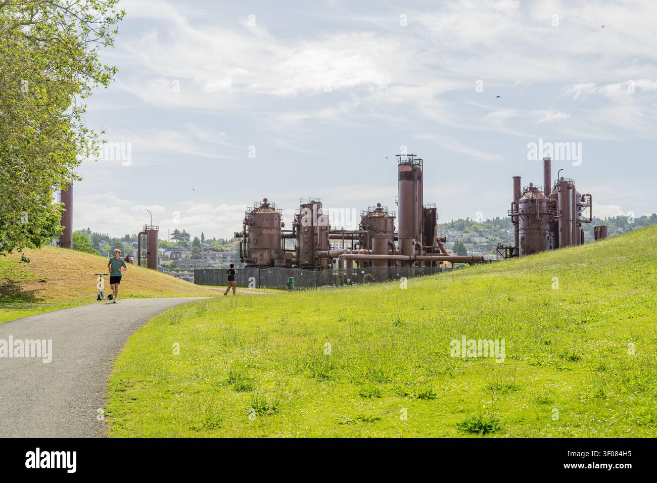 Washington State, JUN 21, 2026 - Verlassene verrostete Industriegebäude und Schornsteine stehen auf einem grasbewachsenen Hügel neben einem gepflasterten Wanderweg mit Stockfoto