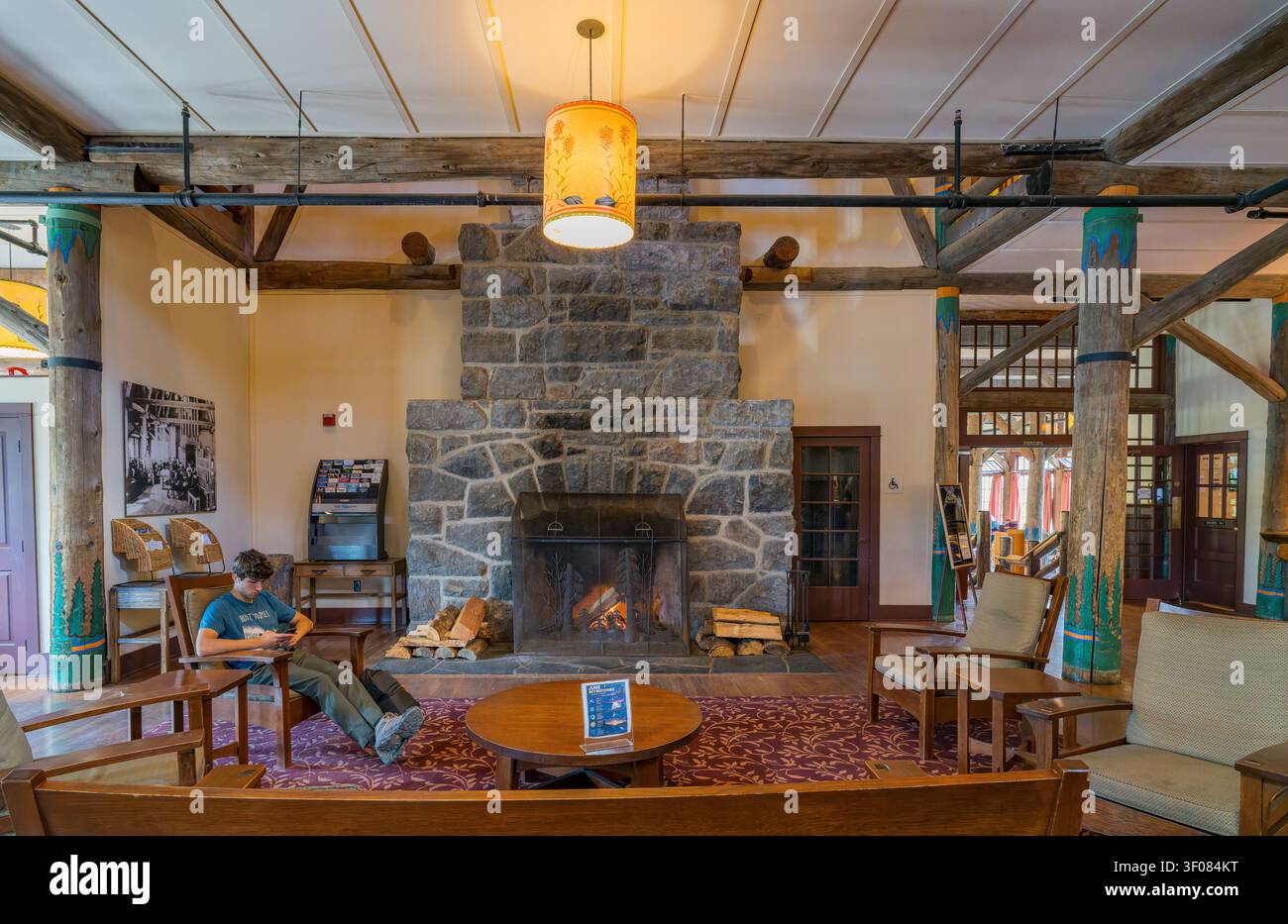 Washington Staat, Vereinigte Staaten, 22. JUN 2026 – Ein junger Mann sitzt in einem Holzstuhl und liest ein Buch in einer rustikalen Lodge-Lobby mit einem großen Stein Stockfoto