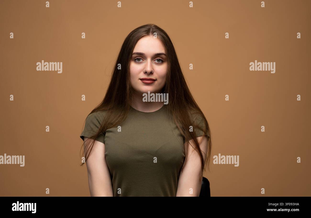 Junge Frau posiert für ein Porträt, zeigt Gelassenheit und Selbstvertrauen mit einem sanften Lächeln auf beigem Studiohintergrund Stockfoto
