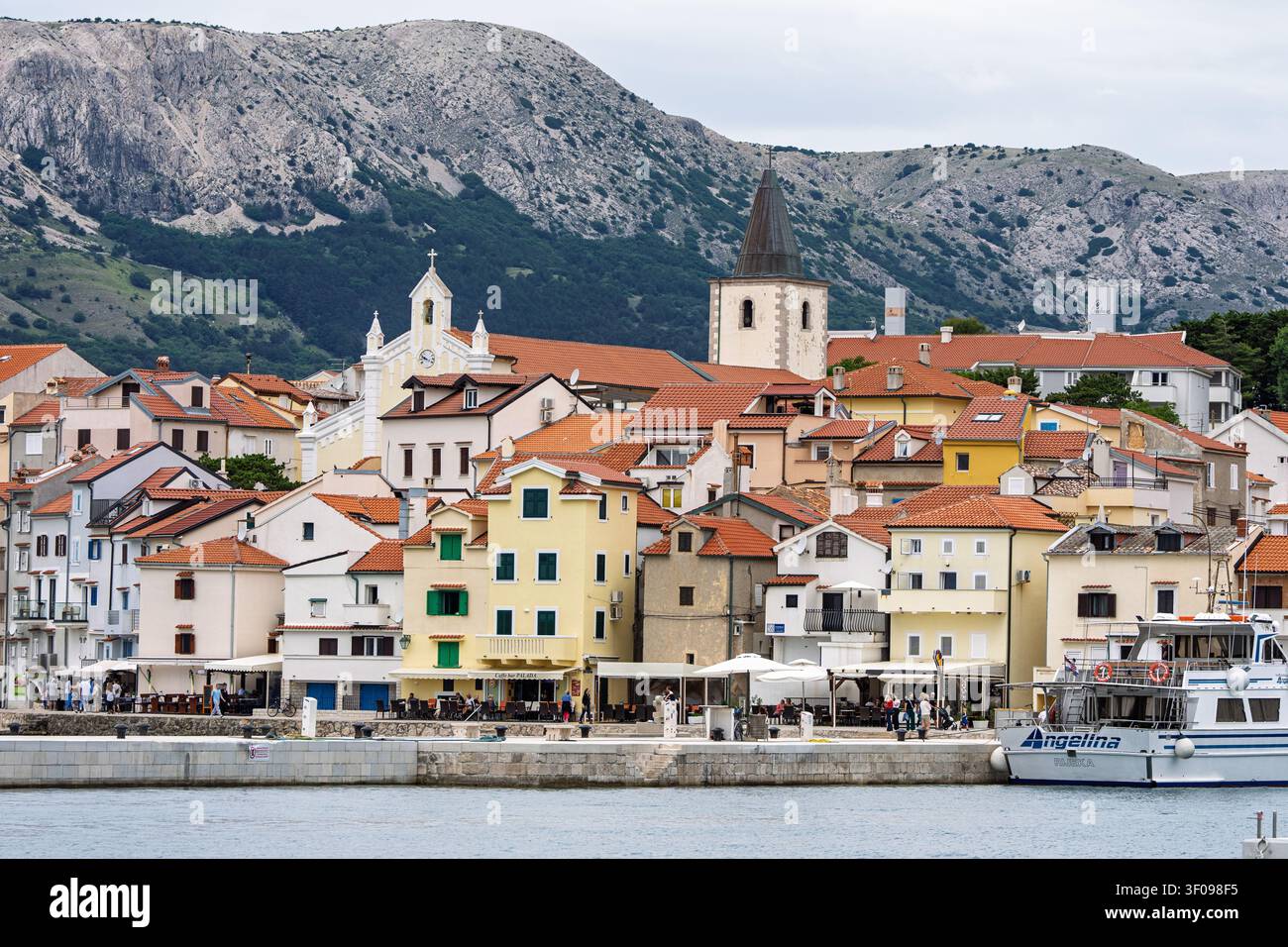 Panoramablick auf die Uferpromenade von Baska mit traditionell roten Ziegeldachhäusern und Hafenpromenade vor felsigen Bergen, mediterranes Küstendorf Stockfoto