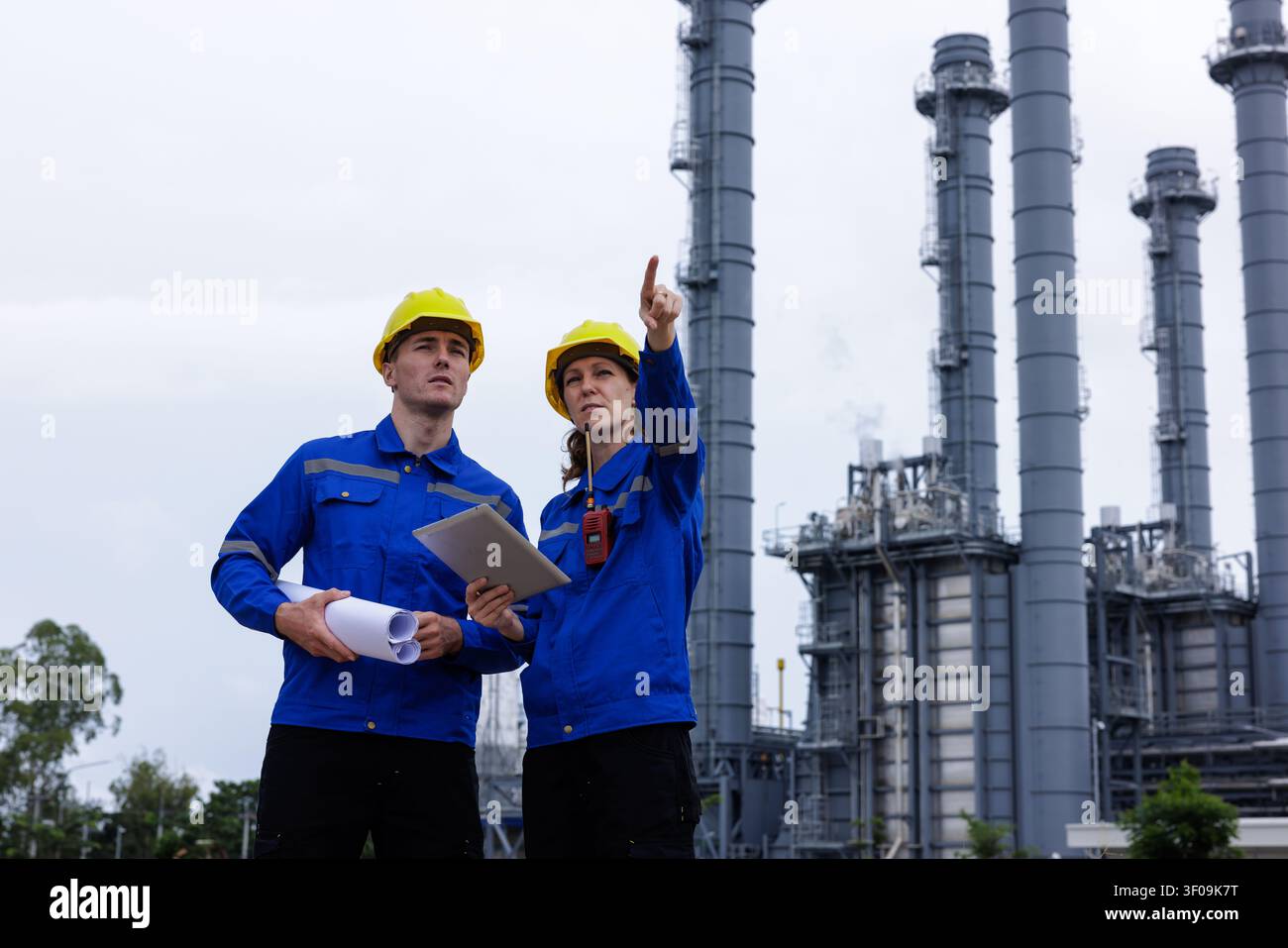 Professionelle Ingenieure in Sicherheitsausrüstung inspizieren eine Energieanlage und überprüfen Infrastrukturpläne auf einem Tablet, um Wartungsarbeiten zu koordinieren. Stockfoto