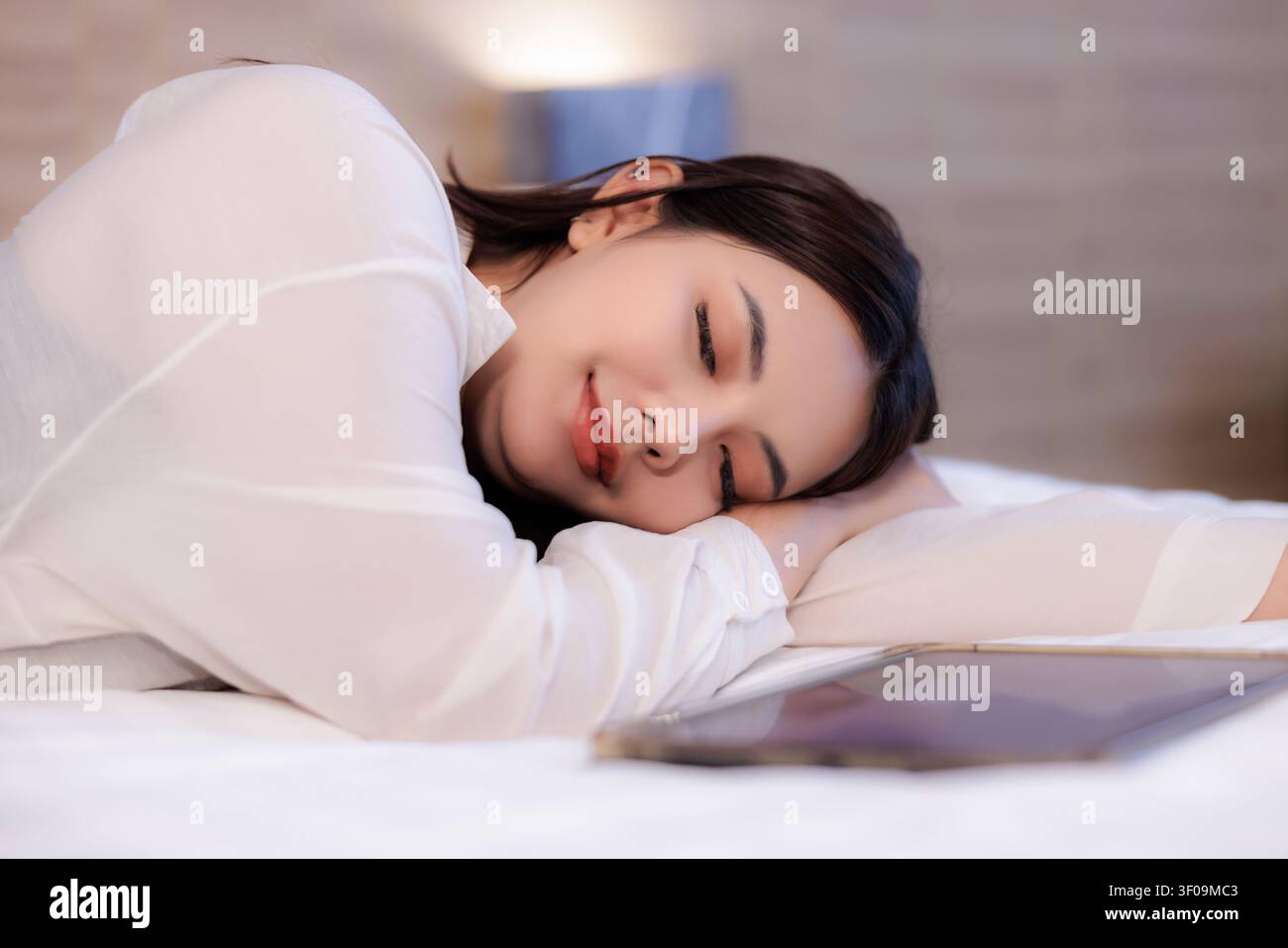 Ein ruhiges Porträt einer jungen Frau, die auf einem Bett neben einem elektronischen Tablet ruht, was einen stillen Moment der Entspannung und Freizeit zu Hause suggeriert. Stockfoto