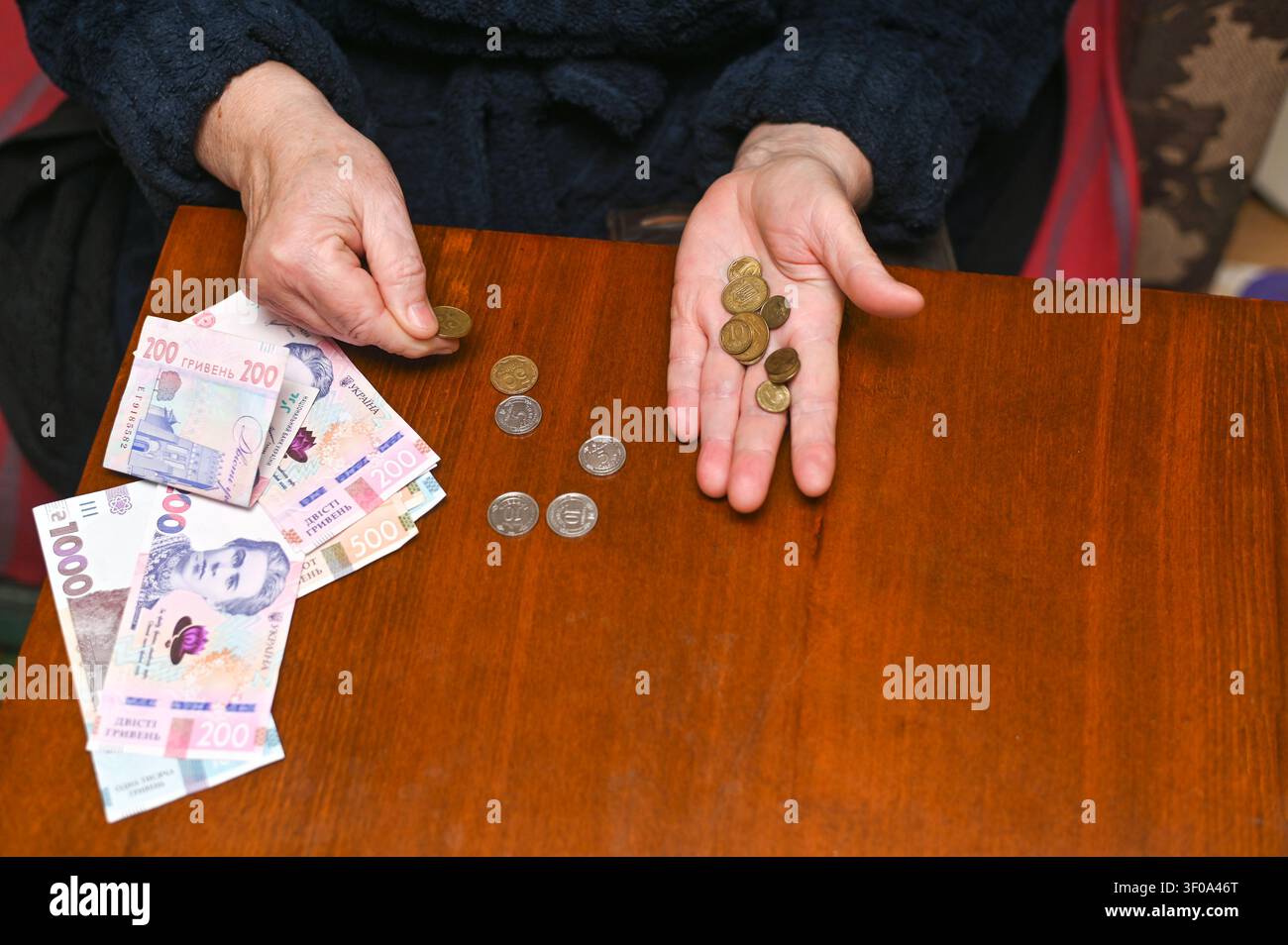 ältere Frau hält kleine Münzen mit Geldbörse, Stapel ukrainischer Banknoten auf Holzoberfläche, Armut und finanzieller Stress, Ältere kämpft mit niedrigem Stockfoto