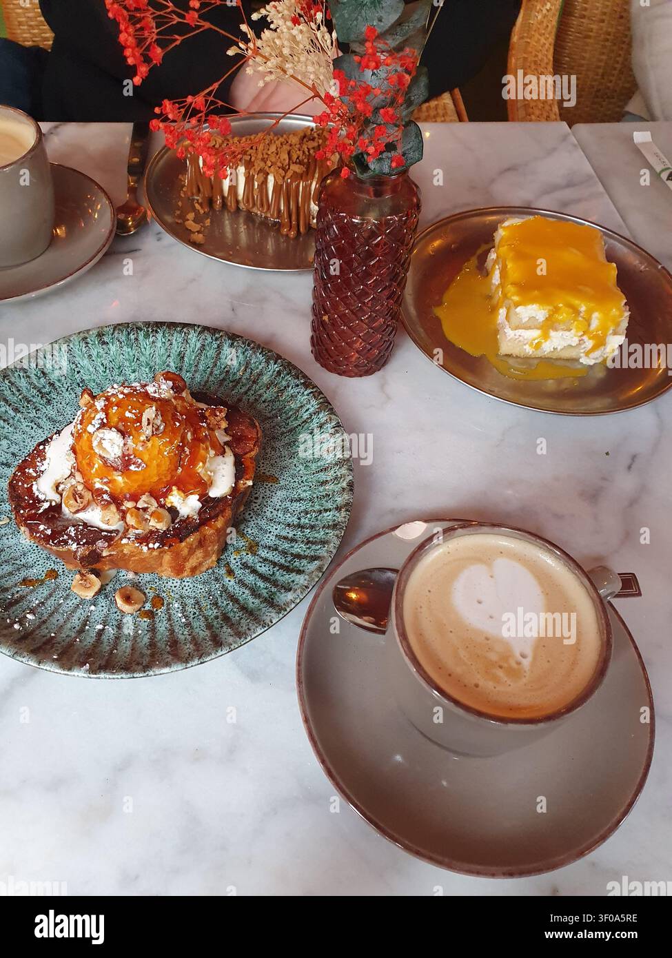 Ein gemütlicher Marmor-Kaffeetisch, der mit einem Latte mit Herzschaum, einem mit Karamell überzogenen Toast-Dessert, einem Mangokuchen auf einem Teller und einer strukturierten Vase mit Dr. arrangiert ist Stockfoto
