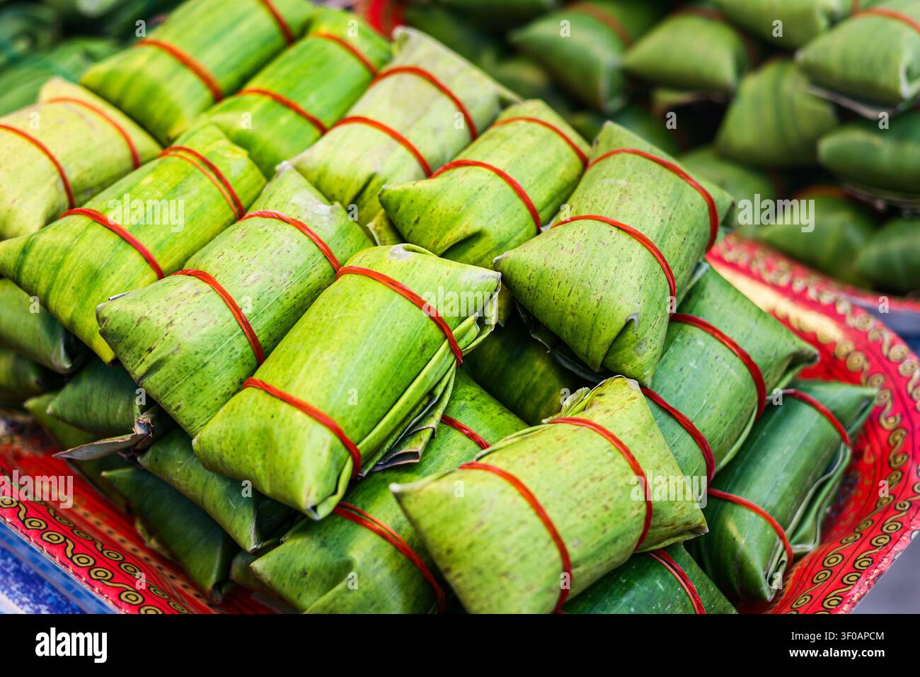 Traditionelle gedämpfte Schweinswurst in frischen grünen Bananenblättern gewickelt und mit roter Schnur gesichert auf einem Tablett auf einem authentischen thailändischen Straßenmarkt. Stockfoto
