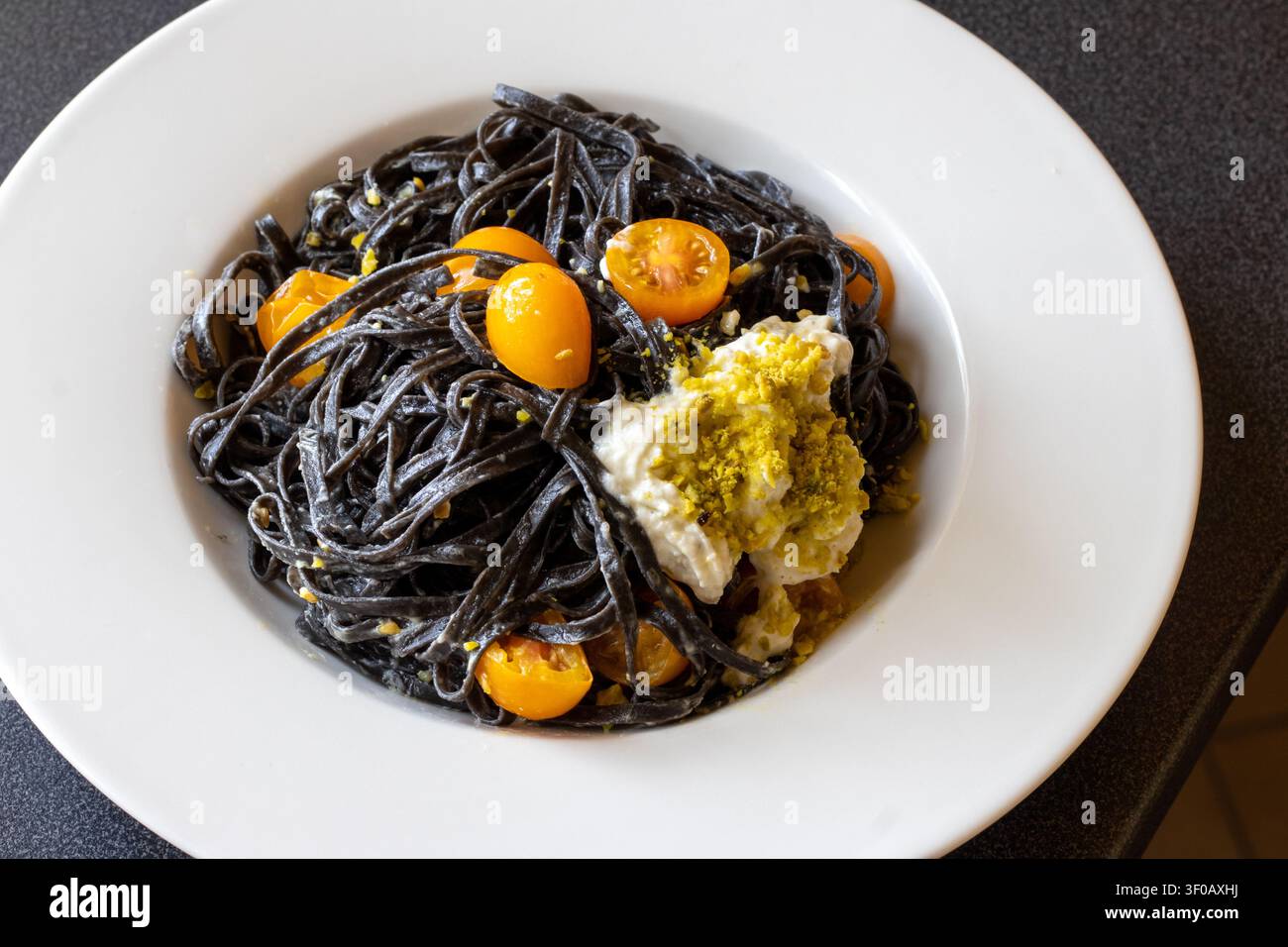 Hochwinkelaufnahme von schwarzen Tintenfisch-Linguine, überzogen mit cremiger Burrata, zerkleinerten Pistazien und halbierten gelben Kirschtomaten in einer weißen Pastaschüssel Stockfoto