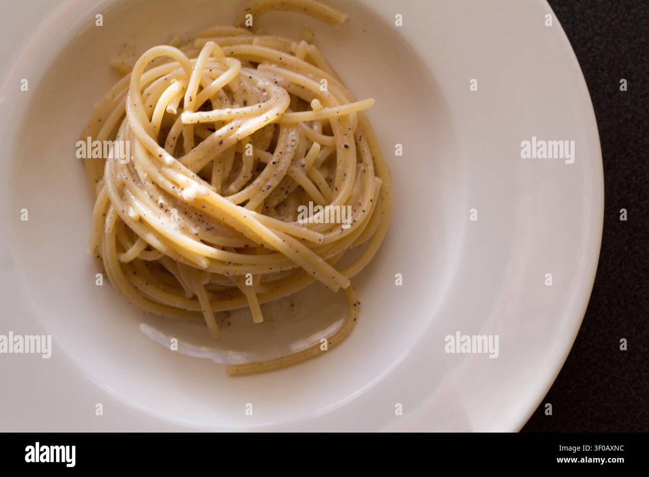 Spaghetti cacio e pepe zu einem Nest geformt mit schwarzem Pfeffer auf einem weißen Teller mit breitem Rand, Ansicht von oben auf einer dunklen Oberfläche, römische Küche Stockfoto
