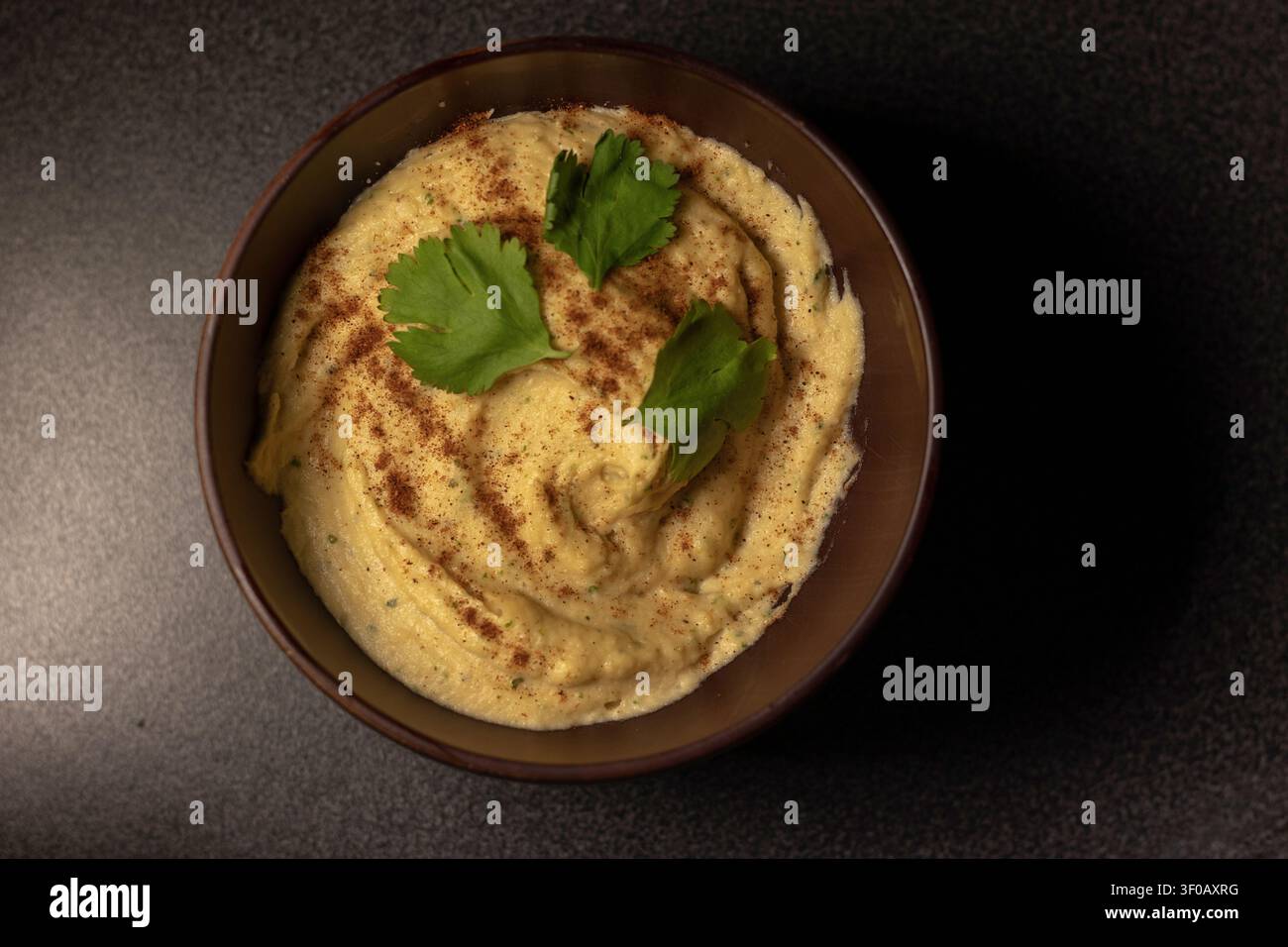 Oberansicht von gewirbeltem Kichererbsen-Hummus mit Paprika und frischen Korianderblättern in einer runden dunklen Schüssel auf einer dunklen Oberfläche, gedämpftes Licht Stockfoto