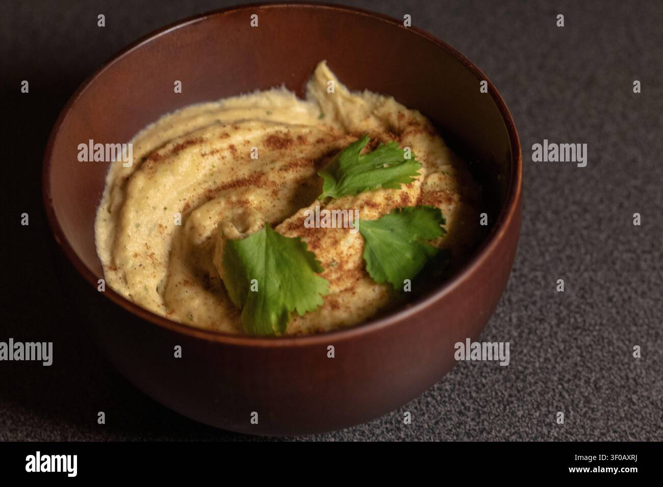 Cremiger, geschwungener Kichererbsen-Hummus mit Paprika und frischen Korianderblättern in einer dunklen Holzschüssel, 45-Grad-Winkel, dunkles und stimmungsvolles Licht Stockfoto