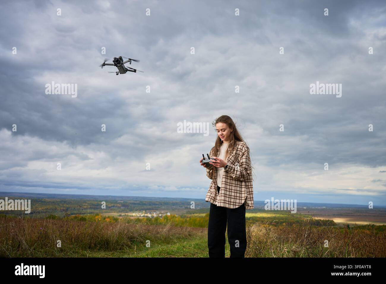 Frau steuert eine Drohne, die am bewölkten Himmel über einer malerischen Landschaft fliegt. Weibliche Pilotin verwendet eine Fernbedienung auf einem Hügel, Konzept der Luftfotografie und Erforschung der Technologie Stockfoto