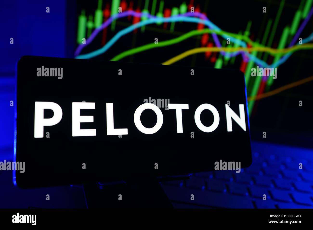 Peloton Interactive Inc-Logo, das auf einem Smartphone-Bildschirm vor einem Computer angezeigt wird, der Aktienmarkt-Finanzleistungsdiagramme zeigt Stockfoto