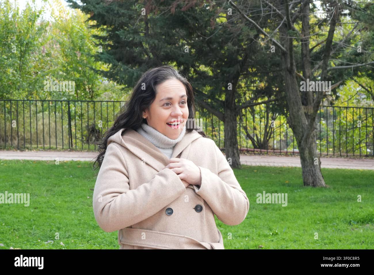Frau lächelt herzlich in einer Parkumgebung. Sie trägt einen hellbeigen Mantel mit hohem Kragen. Ihr dunkles, lockiges Haar fließt sanft um ihre Schultern. Bäume a Stockfoto