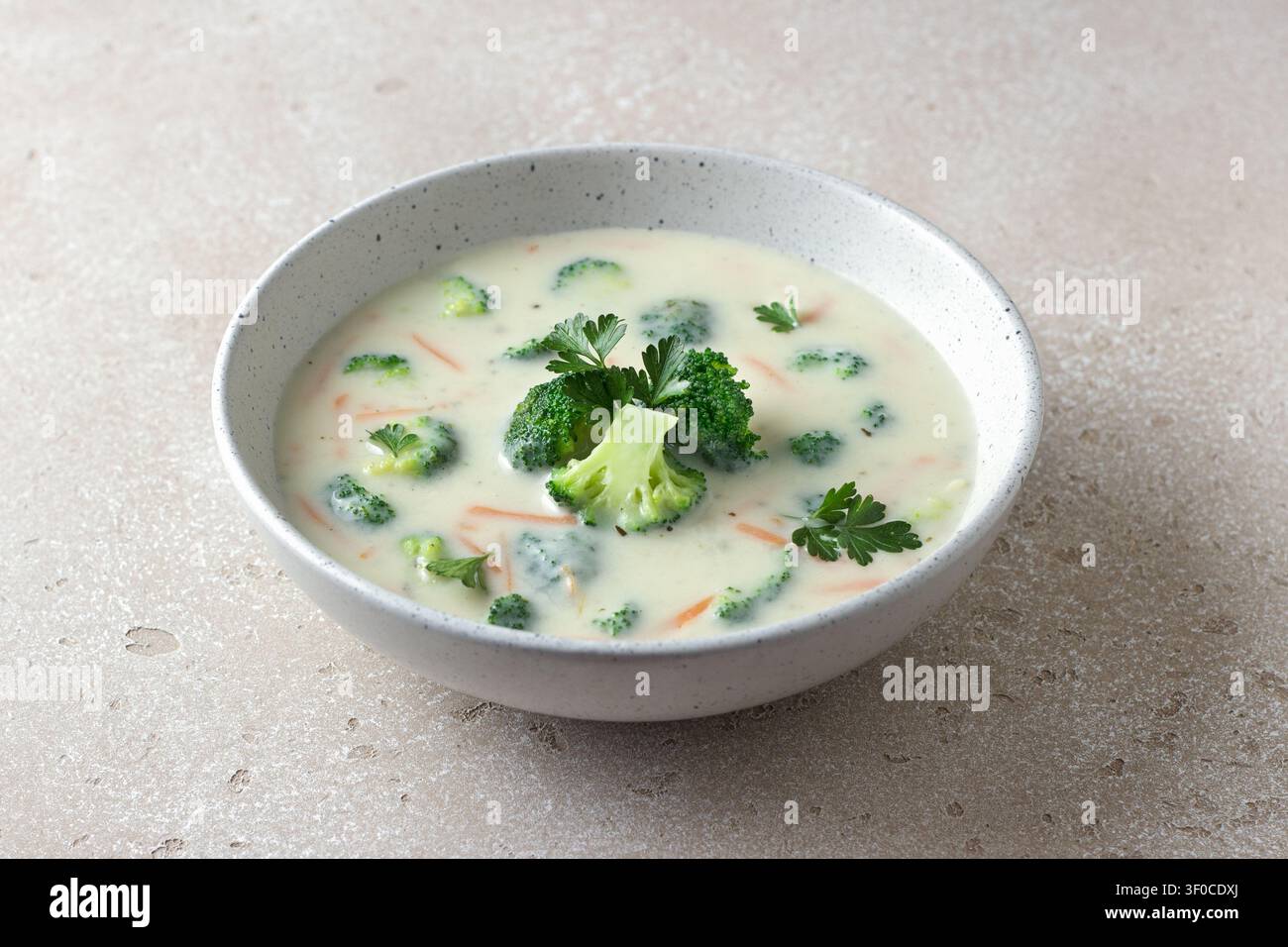 Hausgemachte cremige Brokkolisuppe mit Kartoffeln, Karotten und Petersilie in einer Keramikschüssel auf hellem, texturiertem Hintergrund Stockfoto