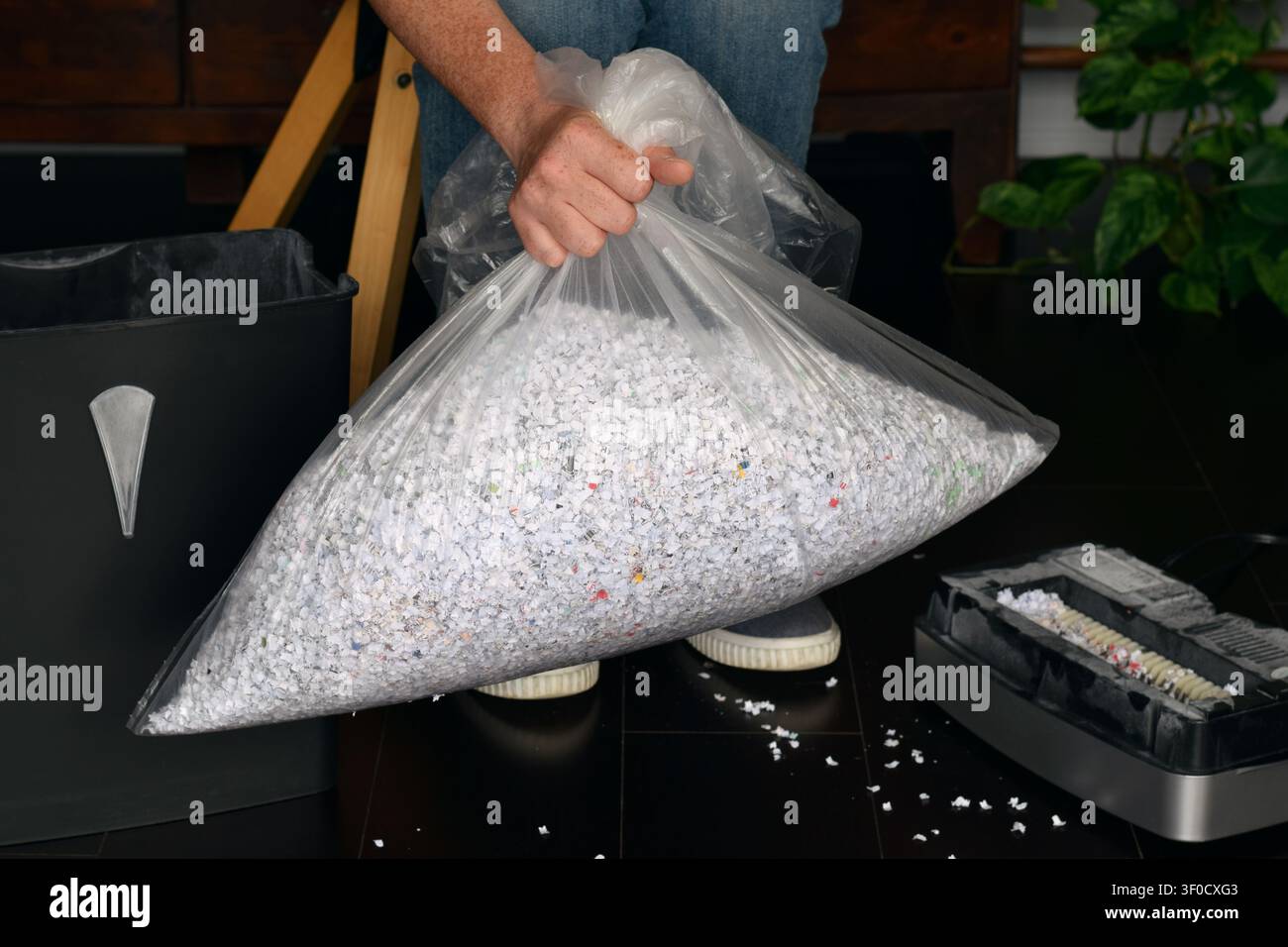 Hochwinkelaufnahme zeigt eine Person, die einen durchsichtigen Plastikbeutel mit geschreddertem Papier neben dem Schredderbehälter und der Motoreinheit auf dem Boden hält Stockfoto